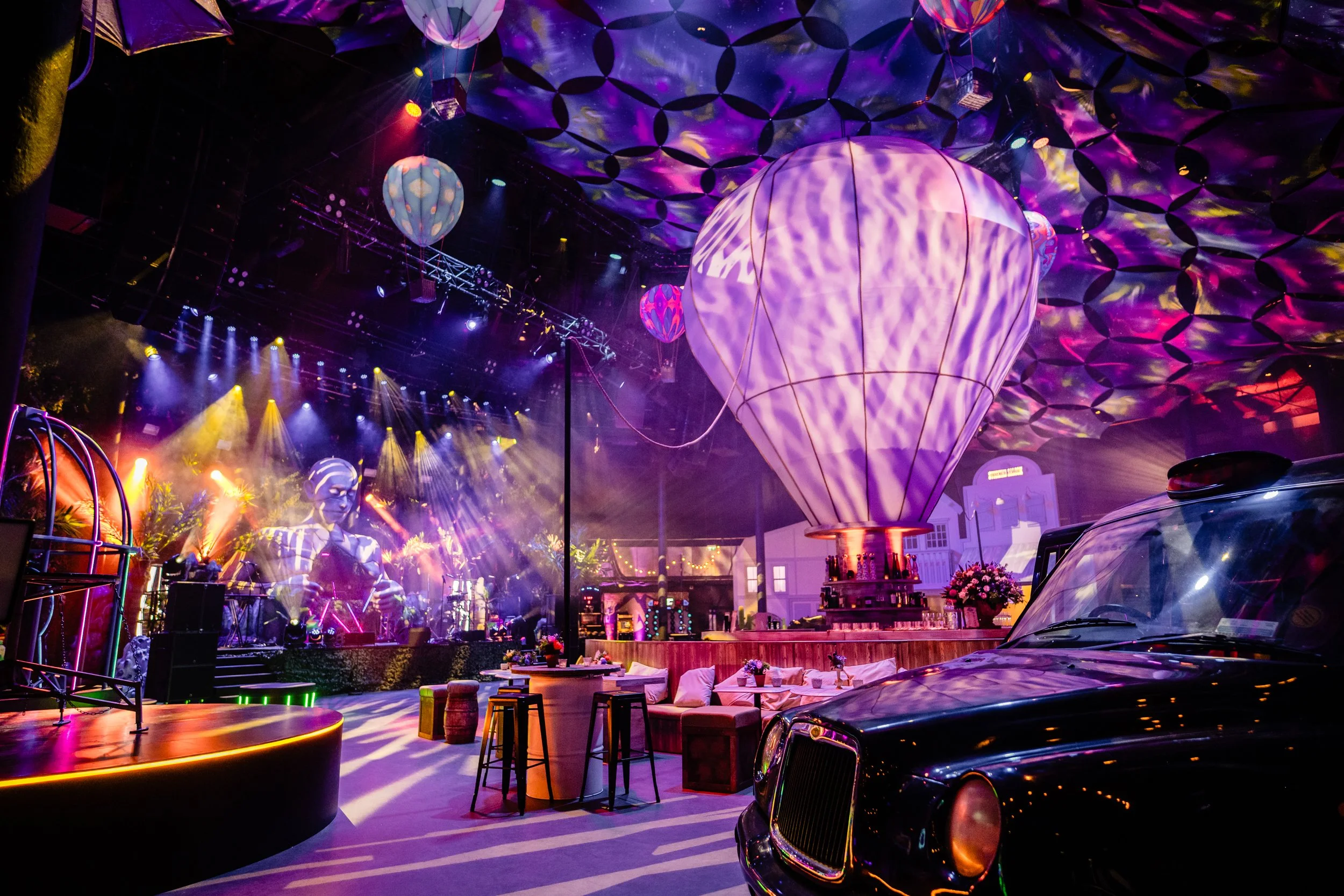 A vibrant Bar Mitzvah event space decorated with colorful lights, hot air balloon lanterns, and artistic ceiling patterns, featuring a stage with a performer, seating areas with flowers, and vintage cars.