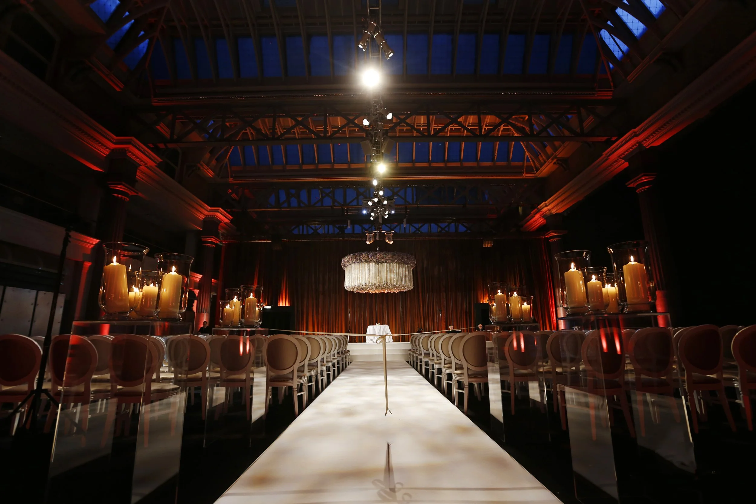 Elegant event venue with a long aisle leading to a stage, surrounded by chairs and topped with large candle arrangements, chandeliers, and warm lighting.