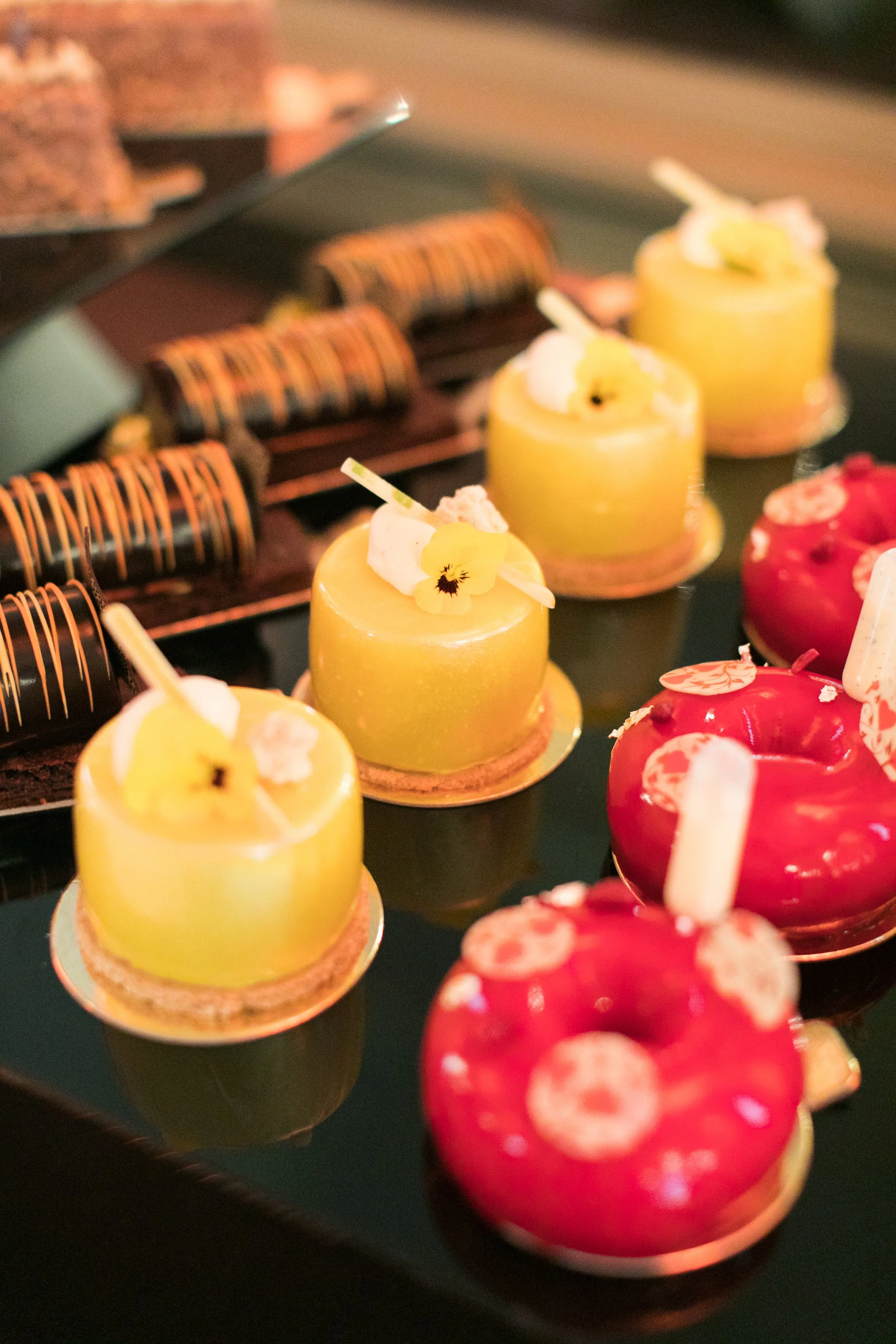Close-up of colorful desserts, including yellow and red glazed pastries topped with edible flowers and whipped cream, arranged on a display tray.