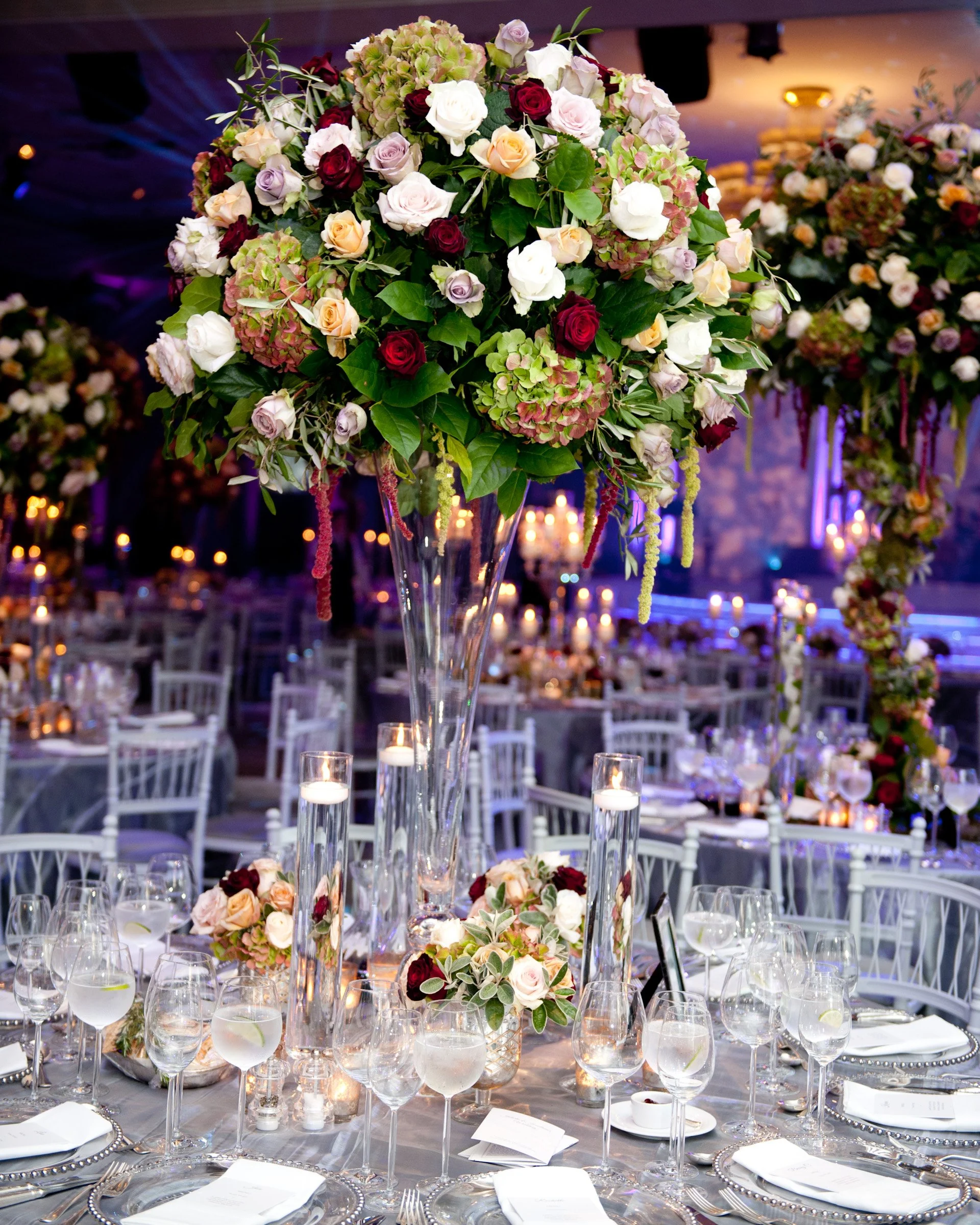 Elegant banquet table decorated with a large floral centerpiece featuring roses and hydrangeas in shades of pink, cream, and burgundy, surrounded by candles, glassware, and white place settings, set for a formal event or wedding reception.