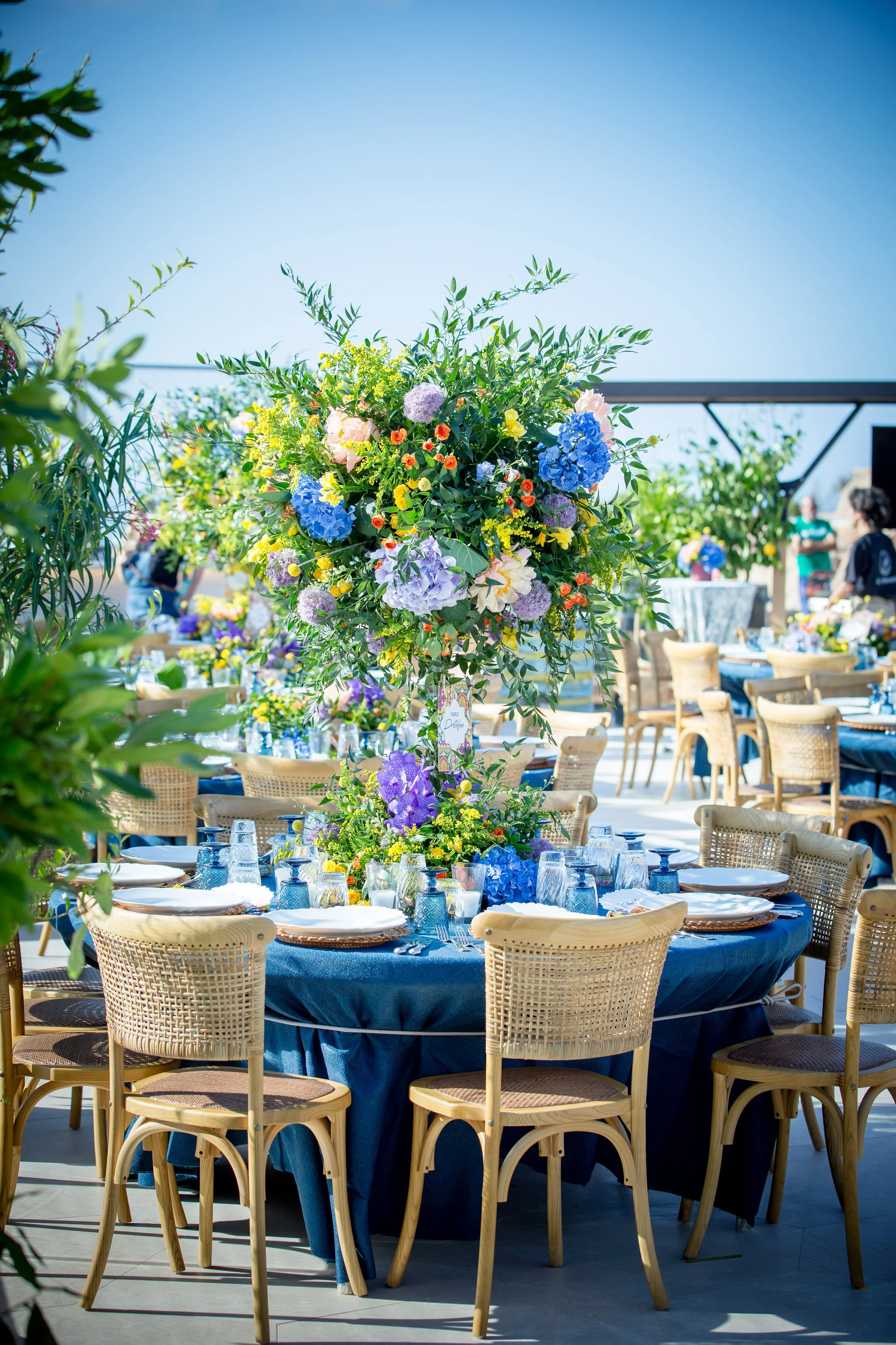 Outdoor event setup with tables draped in dark blue cloth, surrounded by wicker chairs, featuring a tall floral centerpiece with various colorful flowers and greenery under a clear blue sky.