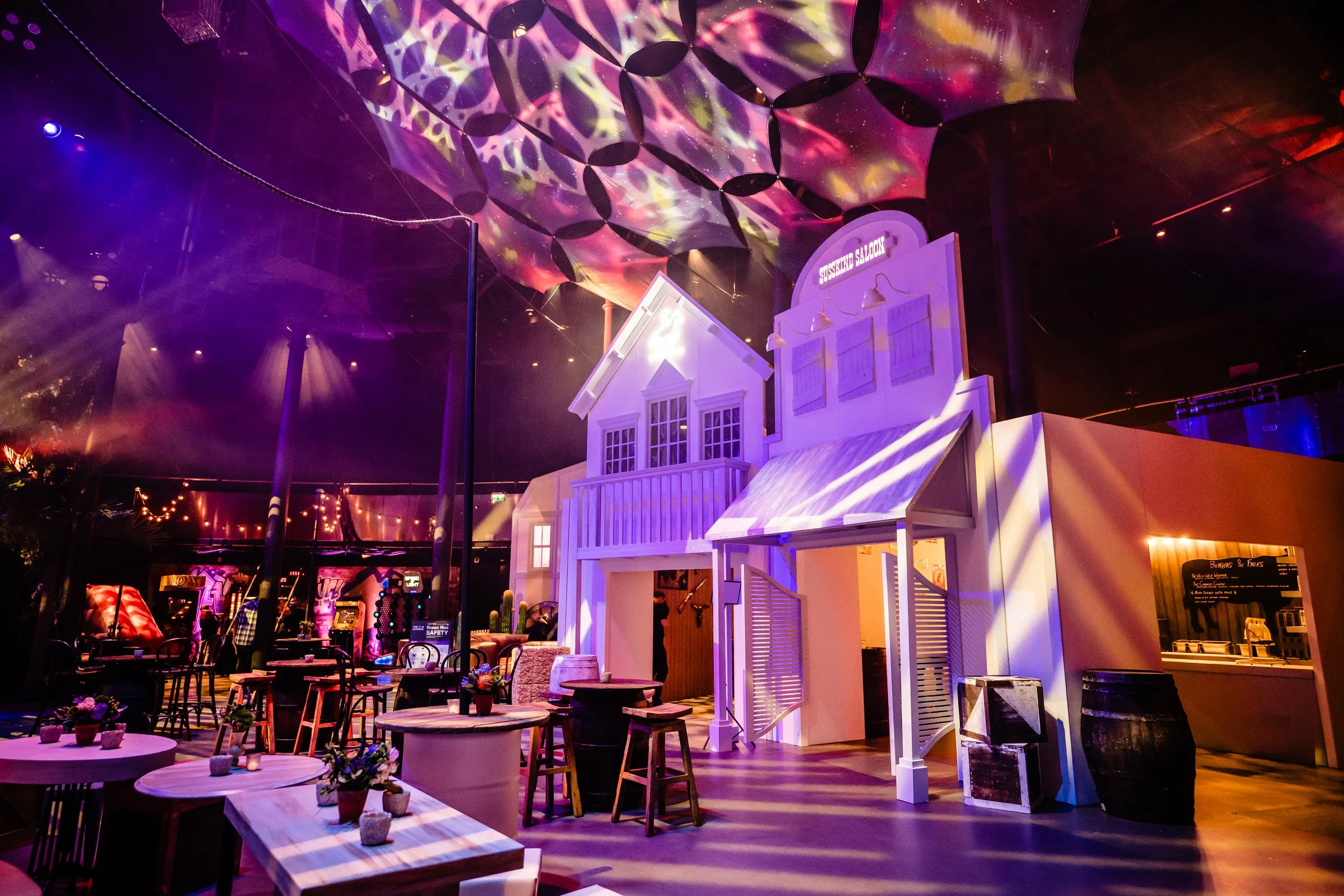 Colorful Bar Mitzvah decorated with pink, purple, and yellow lighting, featuring tables and chairs, and decorative umbrellas hanging from the ceiling.