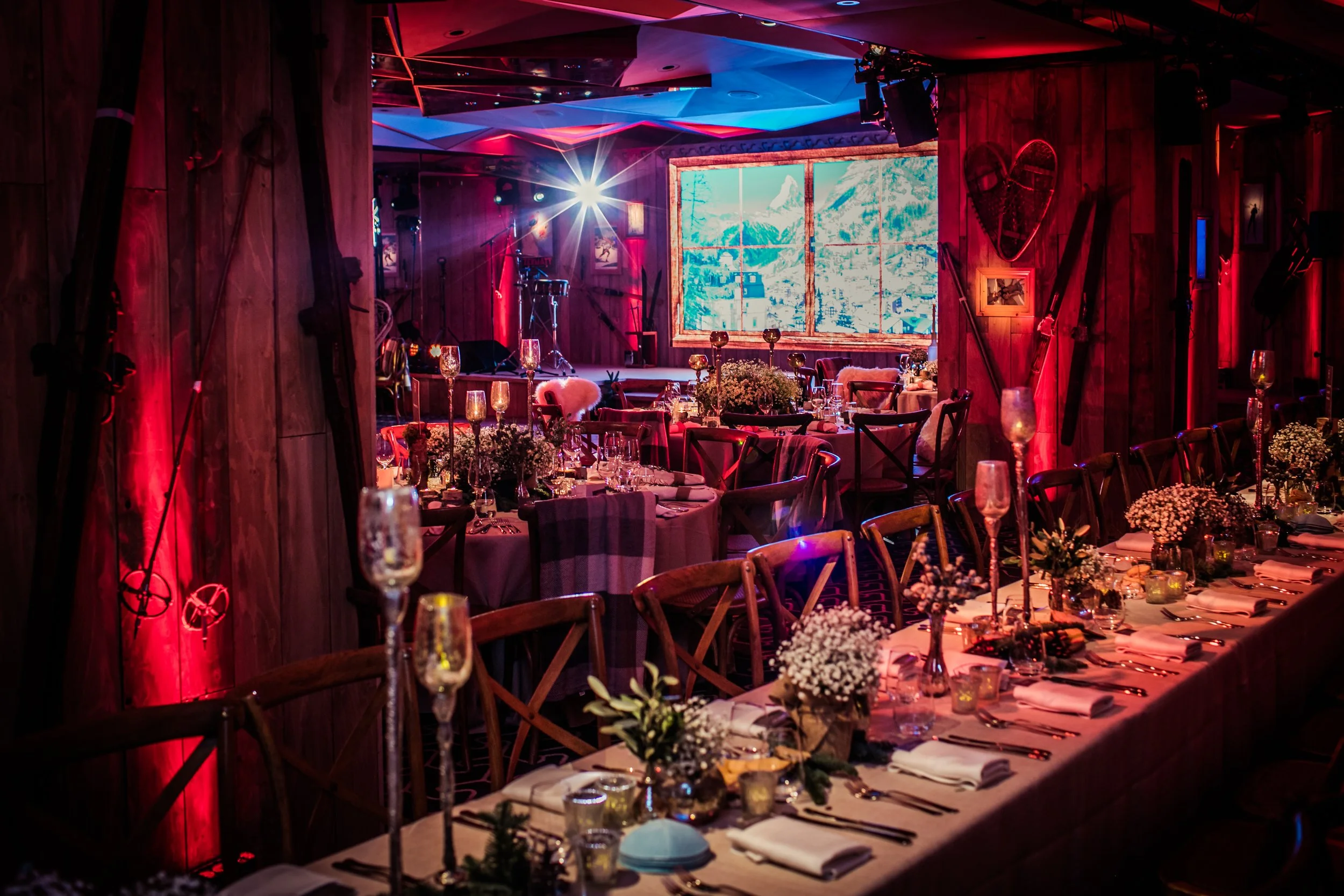 Elegant banquet hall decorated with flowers and candles, set up for a formal dinner with tables, chairs, and a large outdoor mountain scene displayed on a screen.