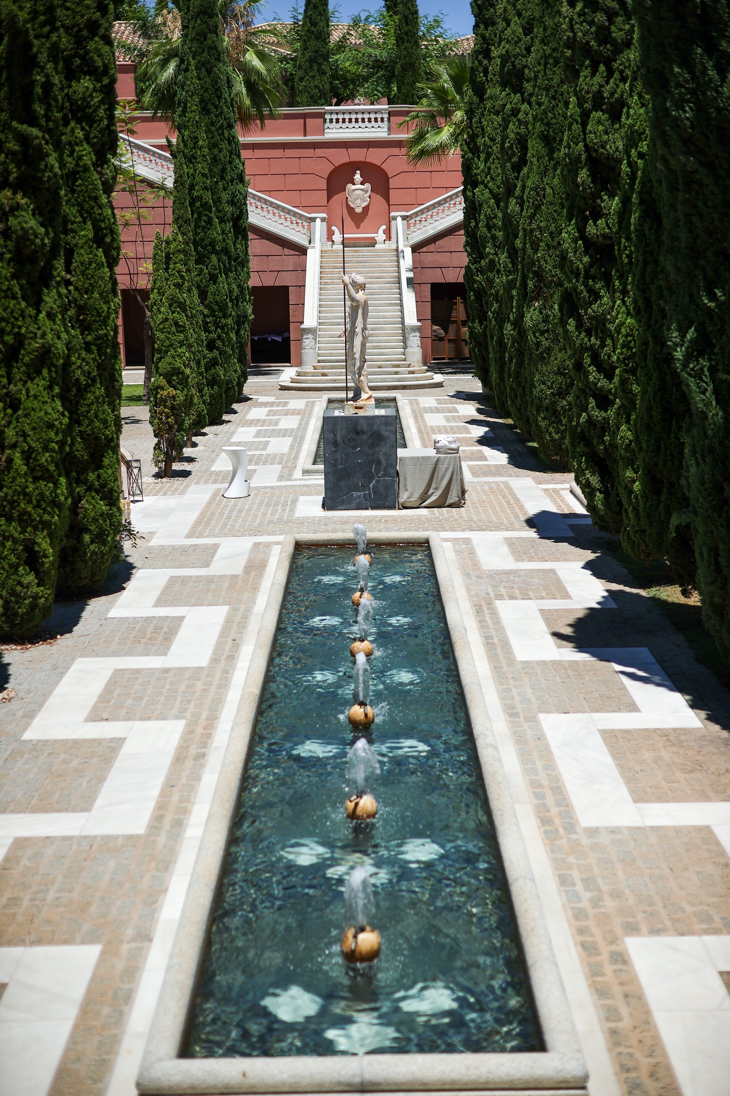 A courtyard with a fountain running through the center, flanked by tall, green cypress trees. There is a marble statue of a woman holding a staff near the fountain. The background features a staircase with white balustrades leading up to a building w
