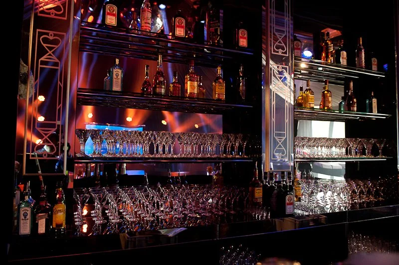 Bar with bottles of alcohol on shelves, many empty glasses, and colorful lighting.