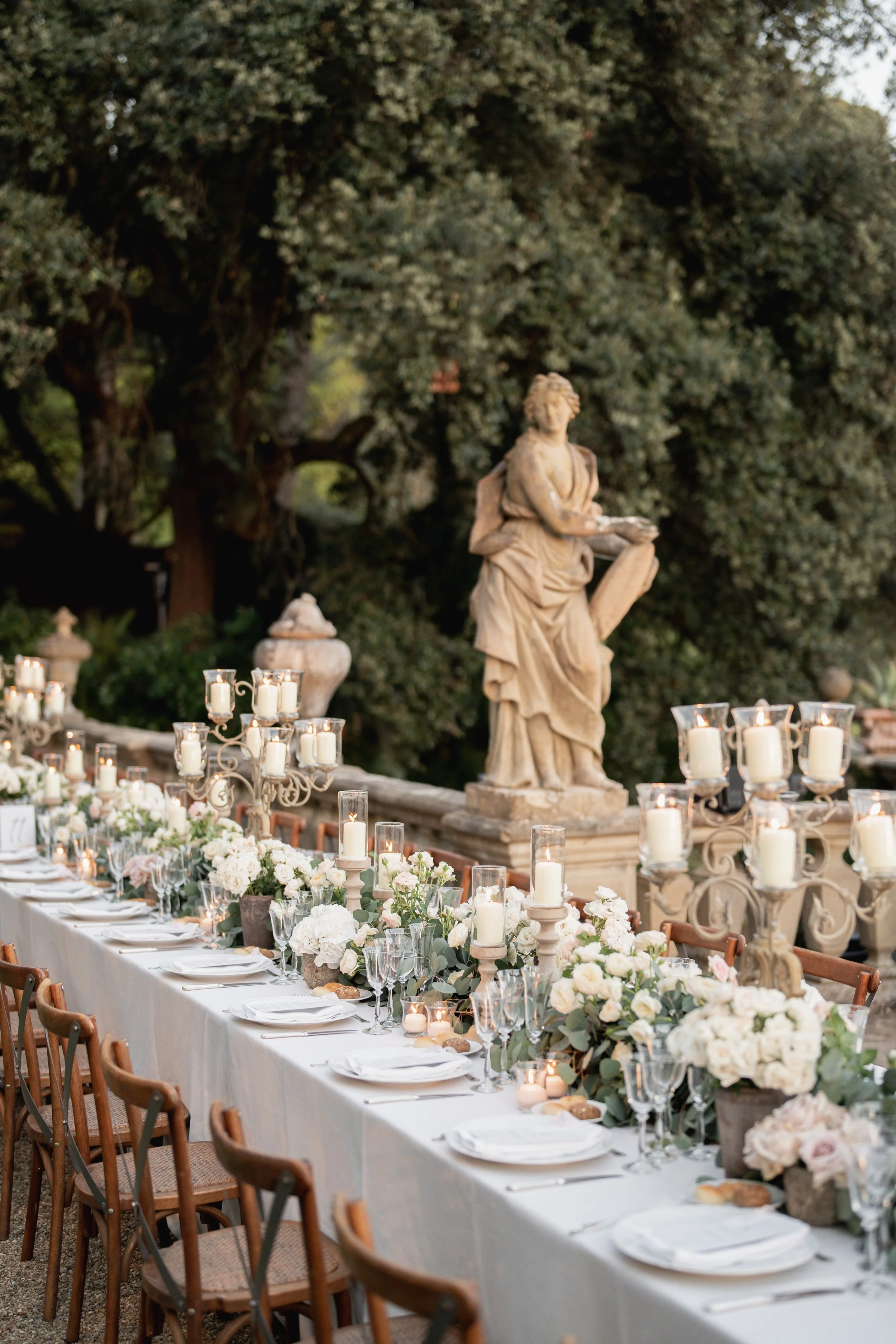 A long banquet table decorated with white flowers, candles, and glassware, set outdoors in front of a large tree and a classical statue at Villa Palmieri in Florence