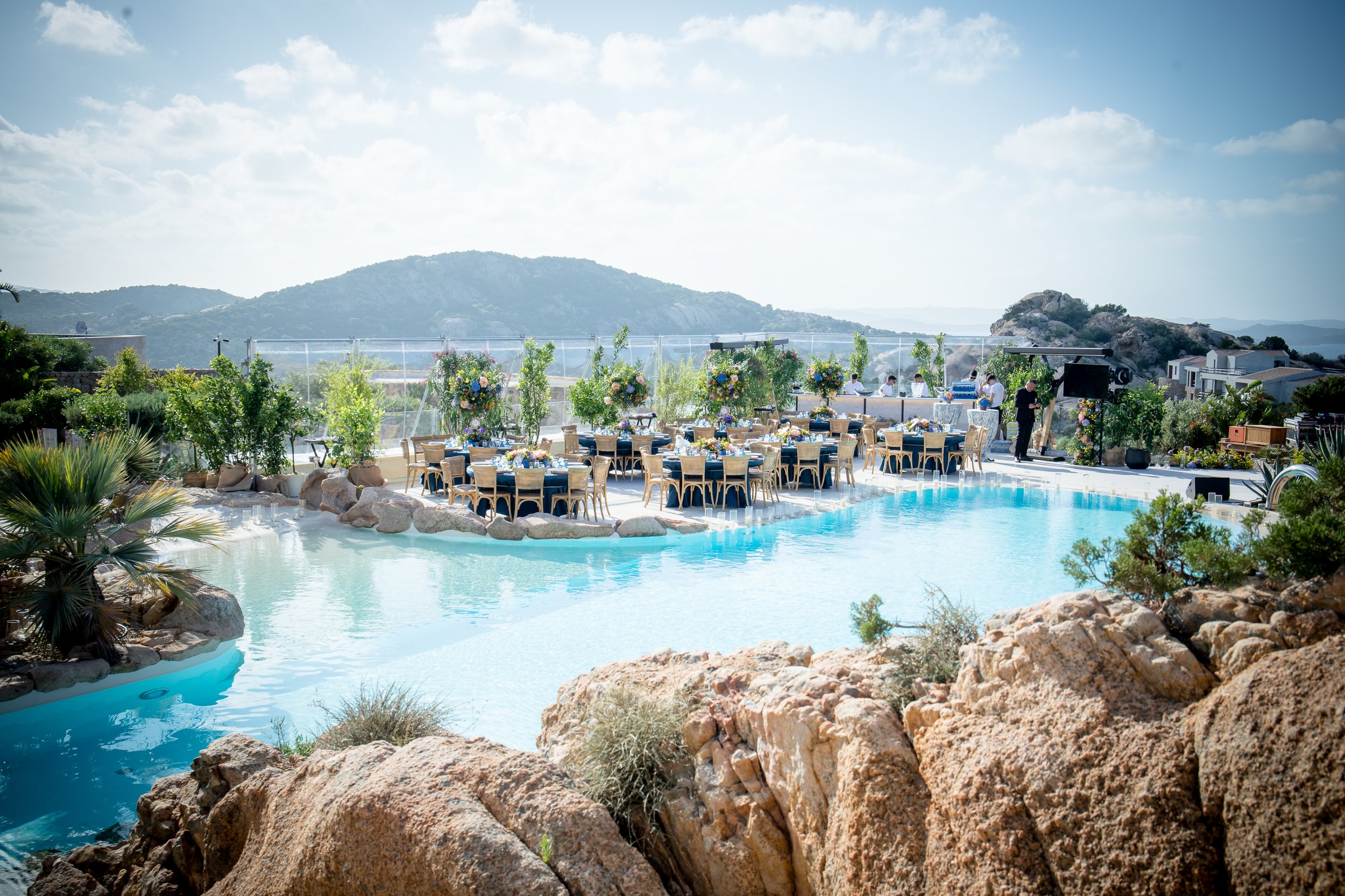 Outdoor event setup at a poolside with tables, chairs, and floral arrangements, surrounded by rocky landscaping with mountains in the background under a partly cloudy sky.