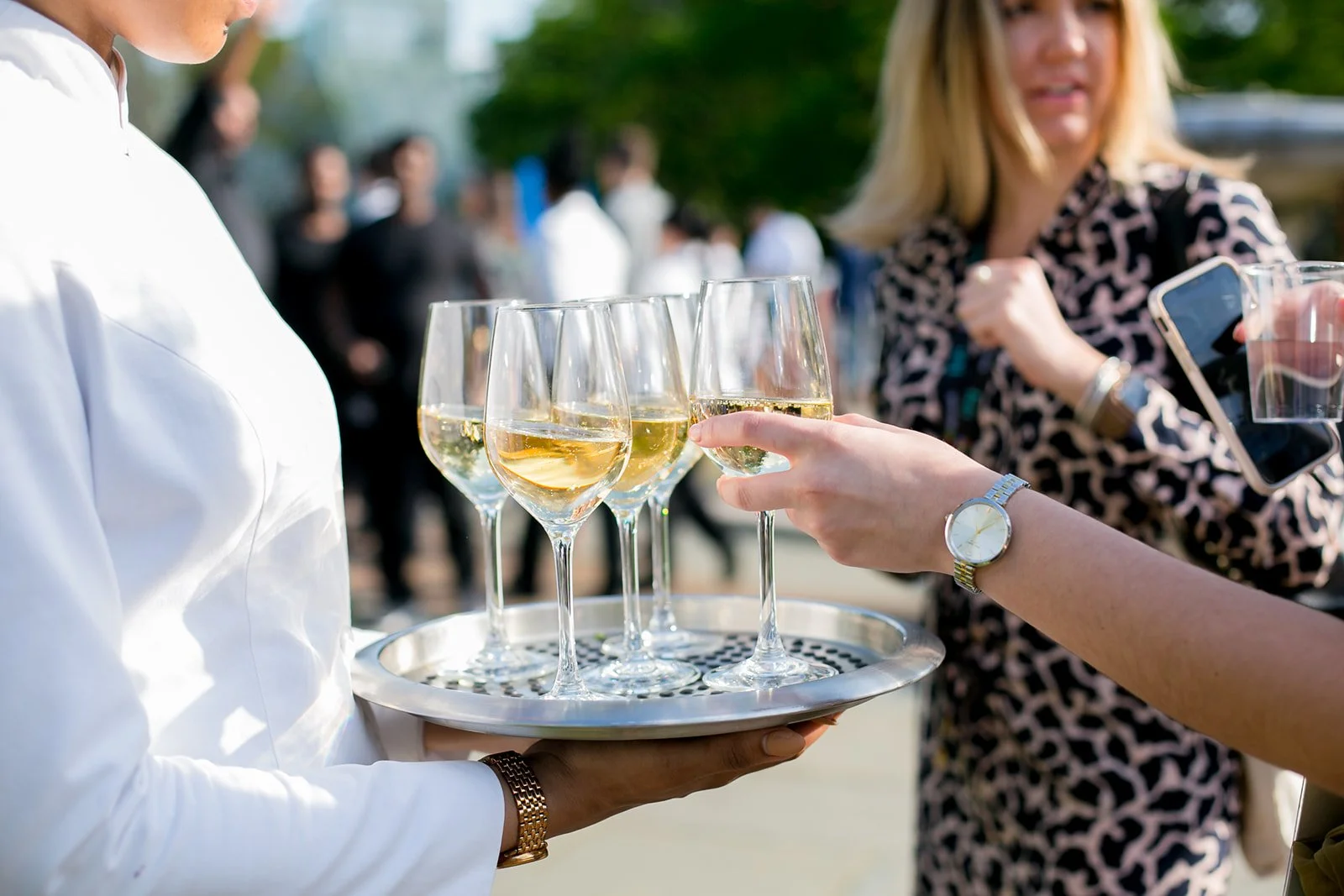 A woman holding a tray of five glasses of white wine while another woman in a leopard print shirt reaches to pick one up. The scene is outdoors with blurred people in the background.