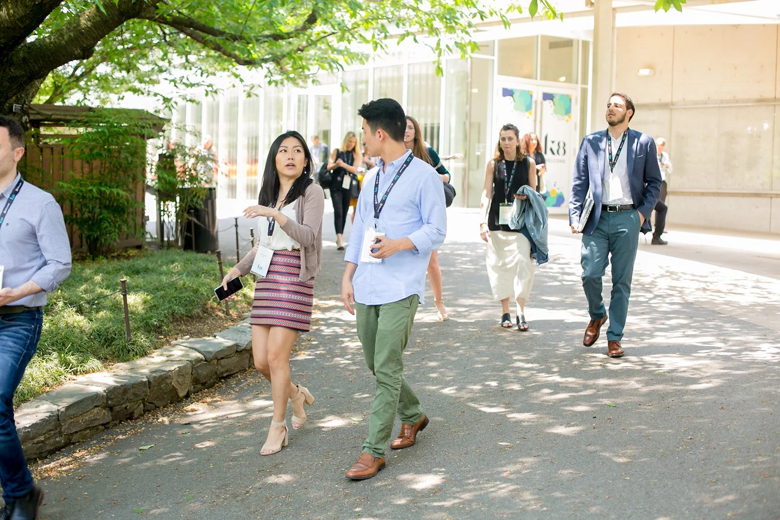 A group of professional people walking outdoors under a tree on a sunny day, engaged in conversation and carrying conference badges.