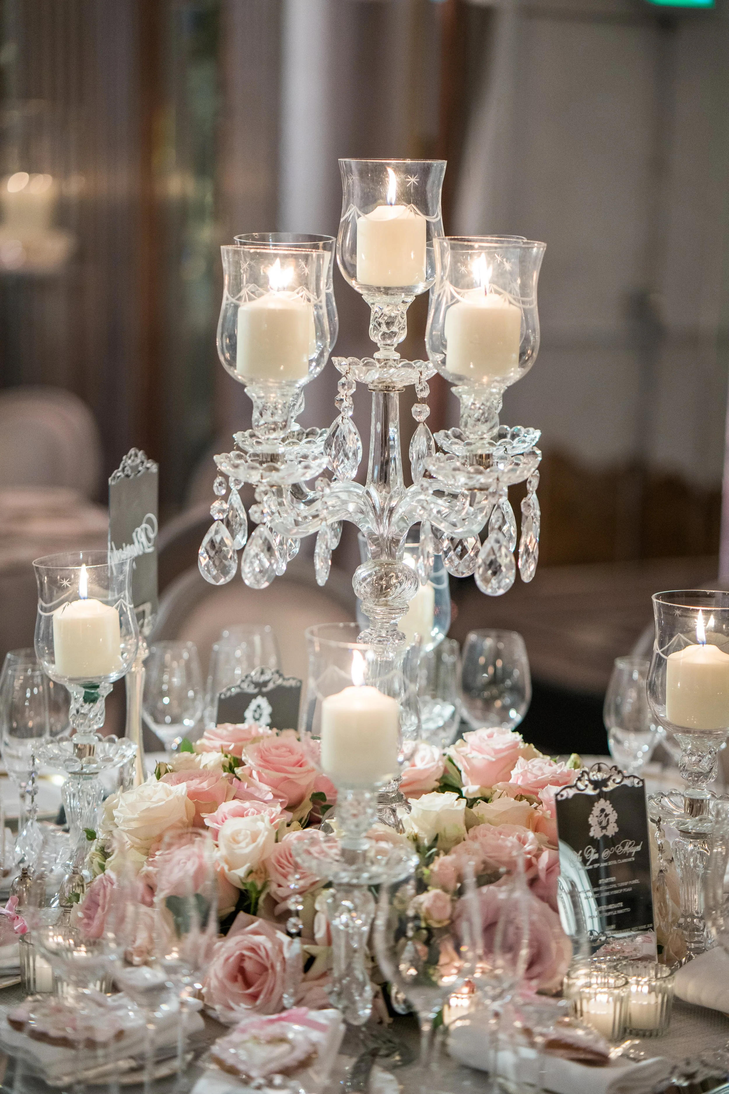 Elegant table centerpiece with a crystal candelabrum holding three lit white candles, surrounded by pink and white roses, and set on a decorated table for a celebration or wedding.