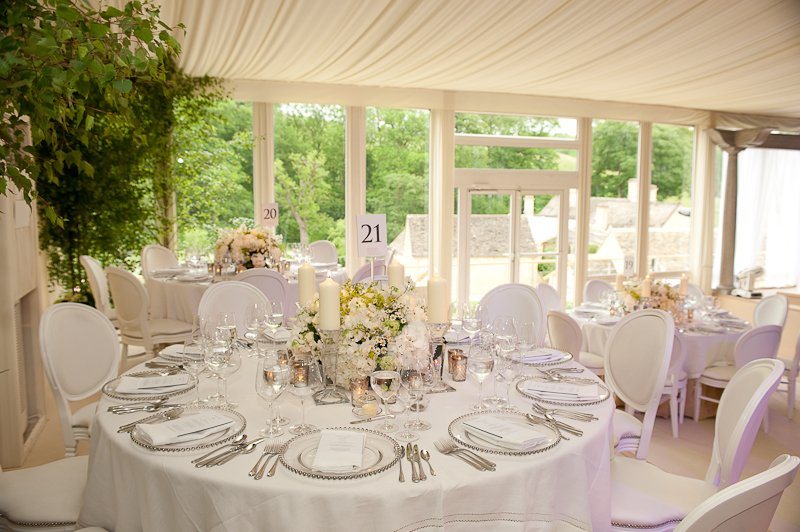 Elegant wedding reception setup with white round tables, floral centerpieces, candles, and white chairs inside a bright tent with large windows and green outdoor scenery.