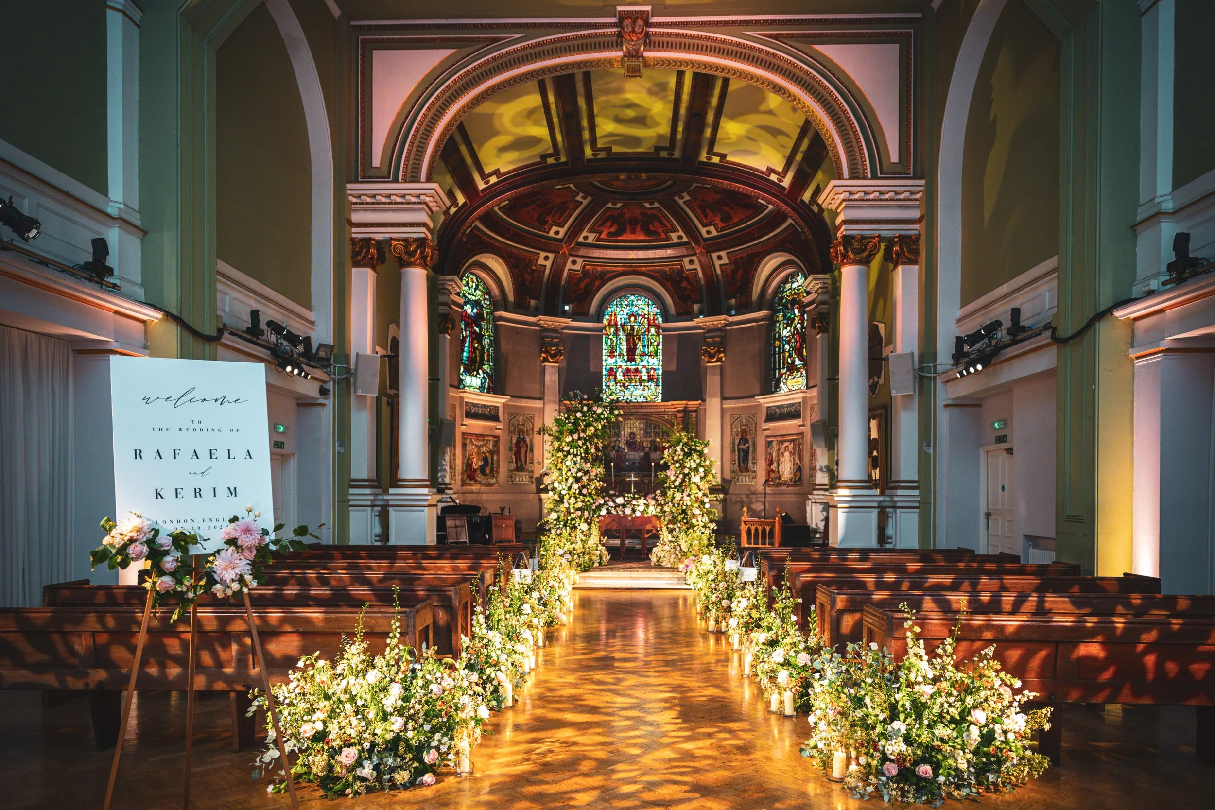 Interior of a church decorated for a wedding, with floral arrangements along the aisle and stained glass windows at the altar.