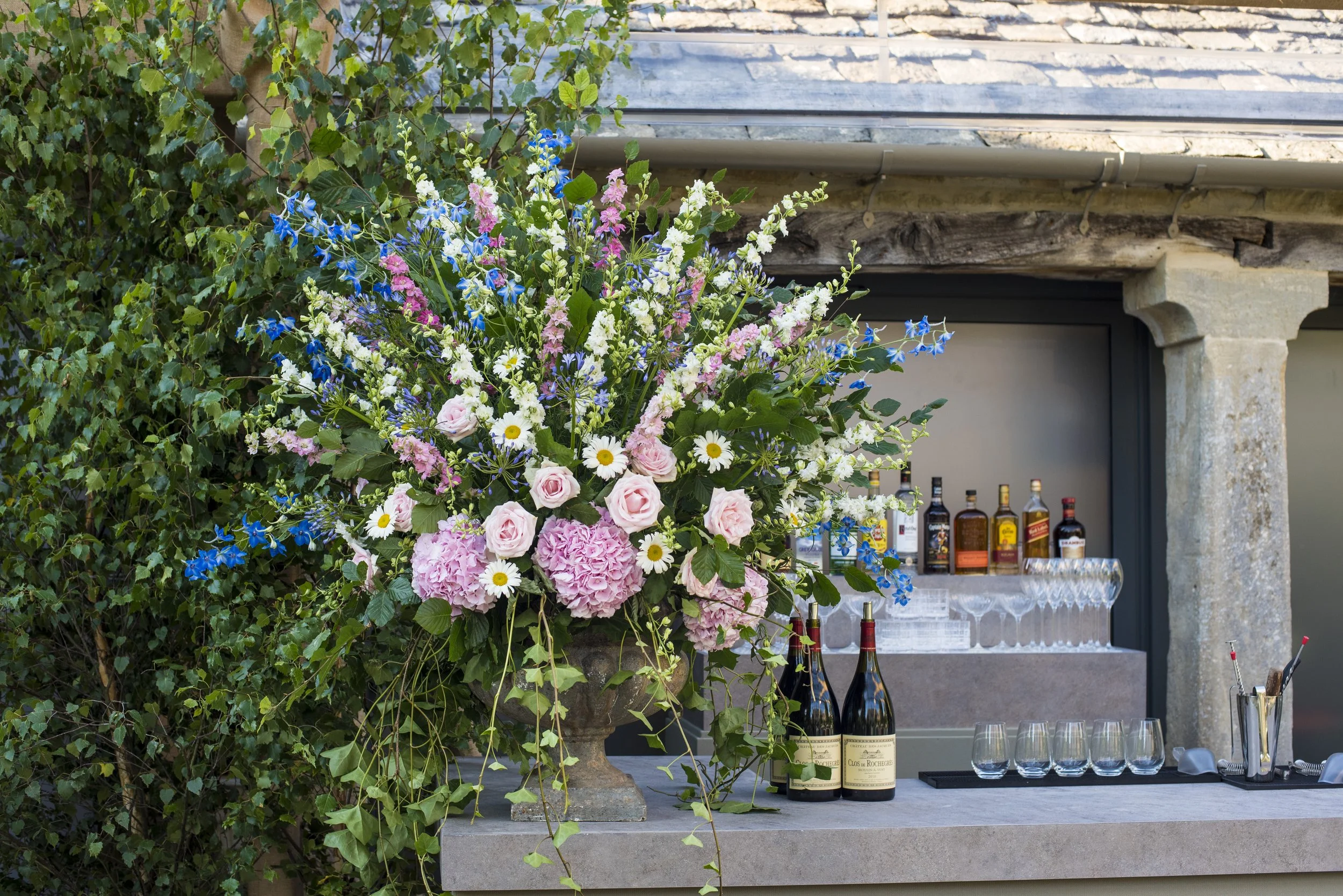 A large bouquet of pink, white, purple, and blue flowers is positioned beside a bar area, with bottles of alcohol and glassware arranged on the bar behind it. The background includes a rustic stone and wood structure.