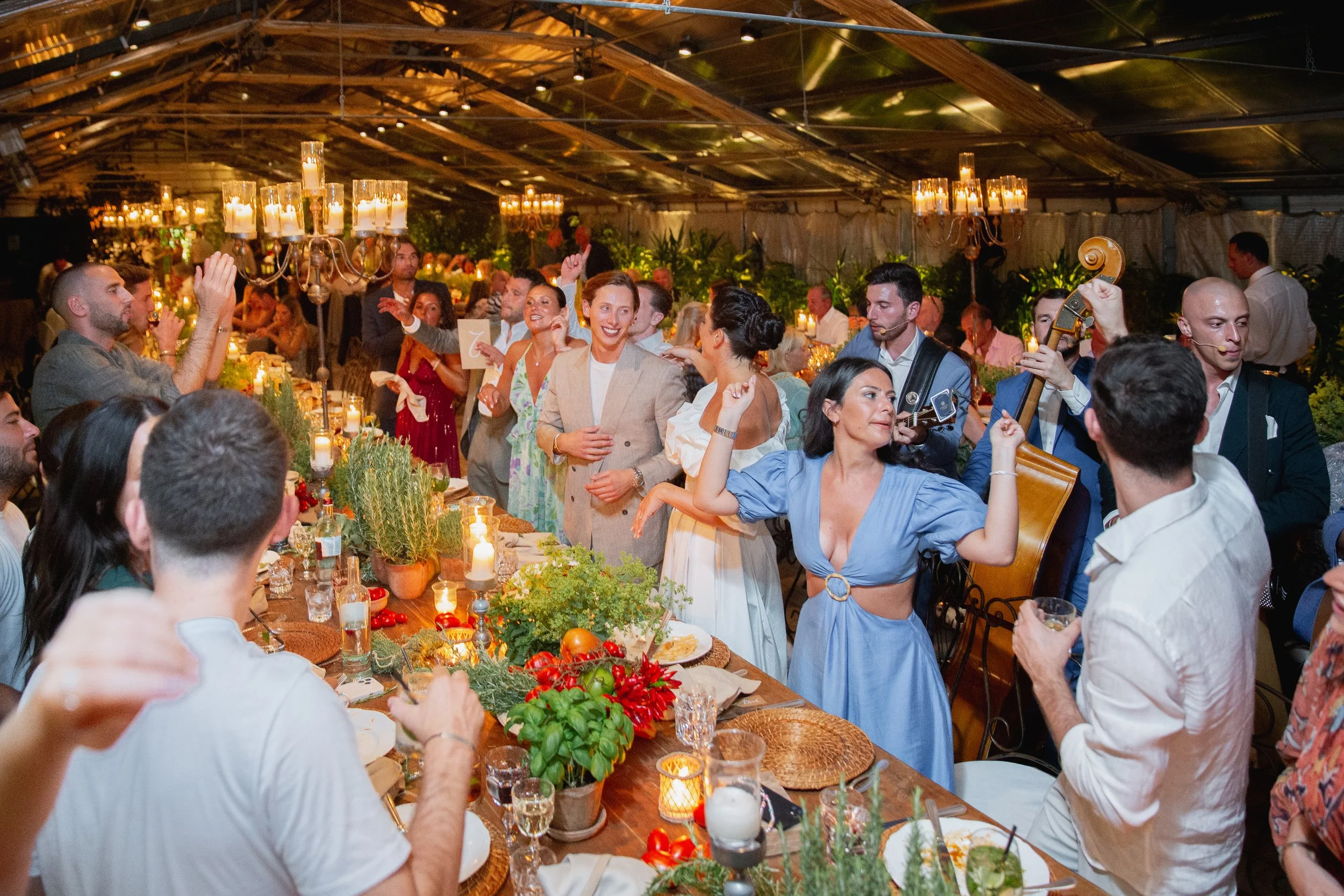 People celebrating at a festive dinner beneath a wooden ceiling, with candles and greenery, some dancing and socializing in Tuscany at Serre Torrigiani Bistrot