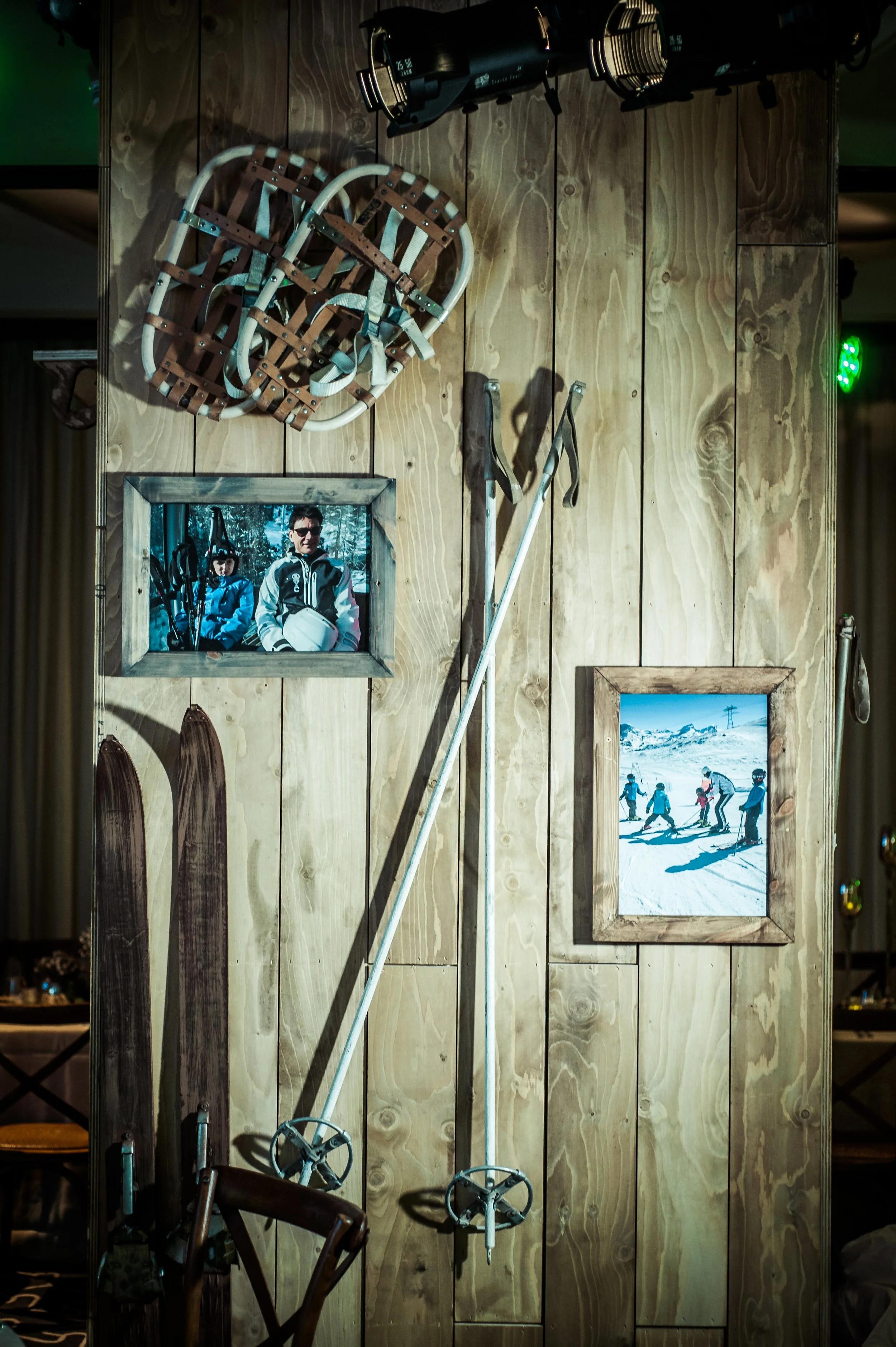 A wooden wall decorated with a framed photo of people skiing, a framed picture of a snowy mountain landscape, skis, ski poles, and ski helmets hanging on hooks, with stage lights above.
