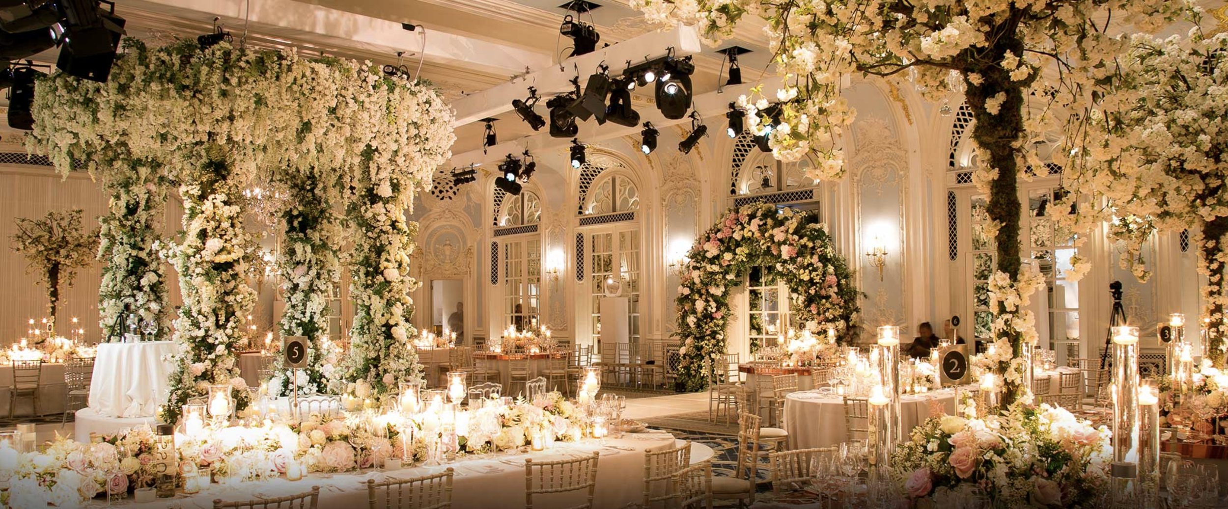 Elegant wedding reception hall decorated with white and blush flowers, including chandeliers, candles, and floral arches, in a grand room with high ceilings and ornate architecture.