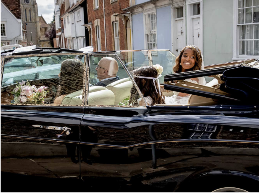 A young woman in a wedding dress sitting in a classic black convertible car, smiling, with two other women and a man inside, on a street with colorful houses and a church in the background.