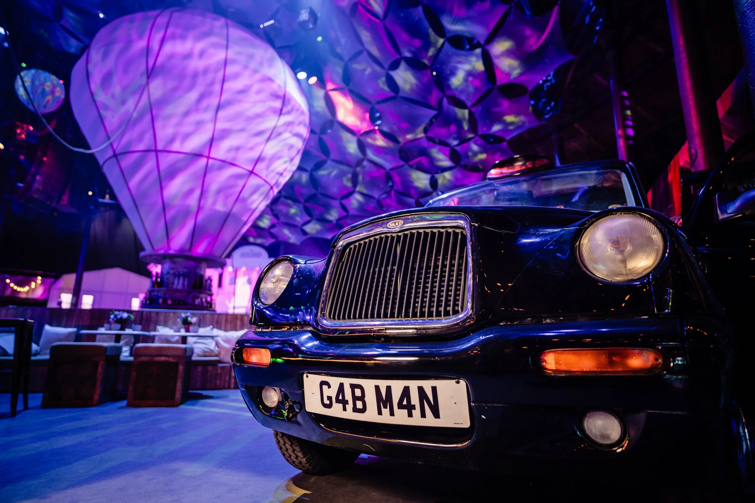 A black vintage car with a license plate reading G4B M4N parked indoors illuminated by purple and pink neon lights. The car's front grille and headlights are visible, with a decorative hot air balloon lamp overhead and colorful ceiling decorations.