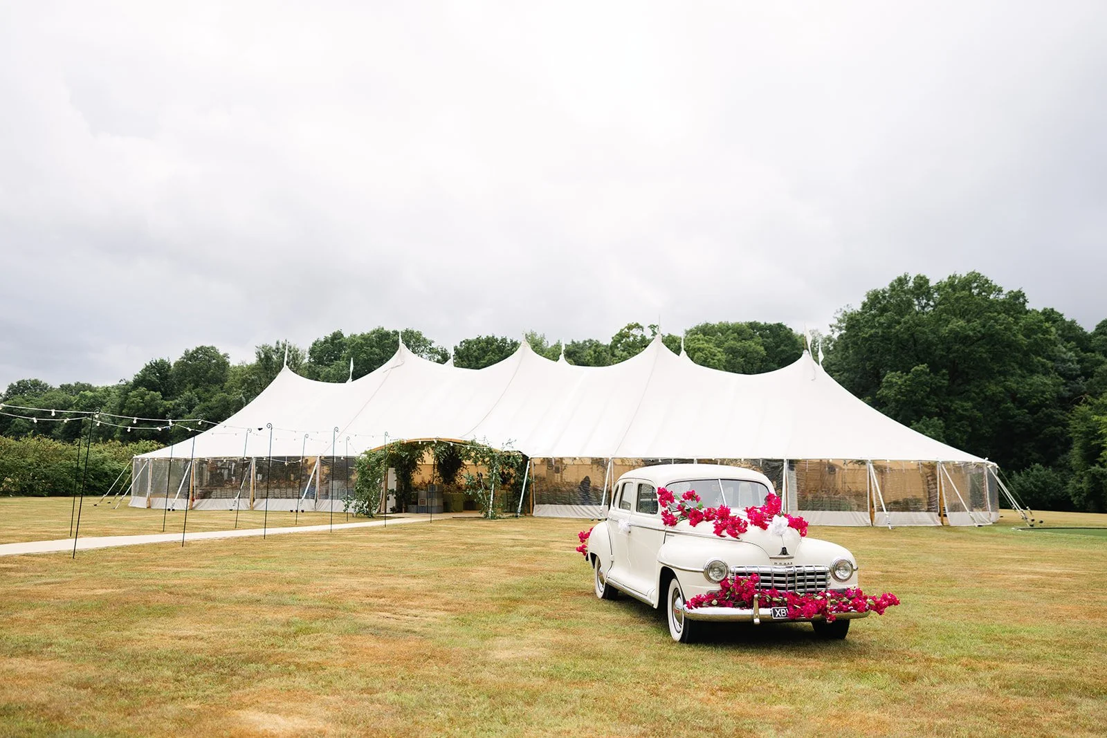 A large white event tent set up on a grassy field with a white vintage car decorated with pink flowers in the foreground, and green trees in the background under a cloudy sky in Hertfordshire