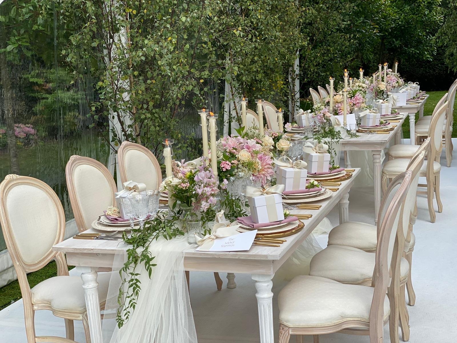 Elegant outdoor dining table decorated with pink and white flowers, candles, and gift boxes, set for a special occasion.