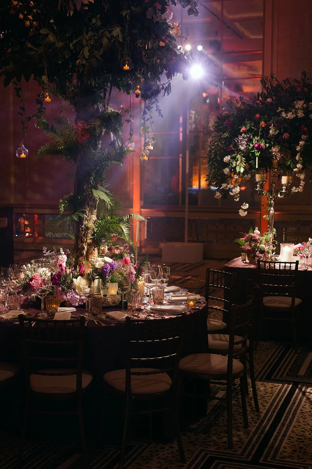 The Peninsula London decorated with floral centerpieces, surrounded by black chairs. Large floral arrangements and hanging lanterns adorn a tree, with candles on the table. Soft, warm lighting creates a romantic ambiance. 