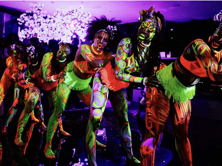 Group of people with body paint glowing under black light, wearing colorful costumes with neon patterns and accessories, performing or dancing at an indoor event with a purple-lit background and a white illuminated tree.