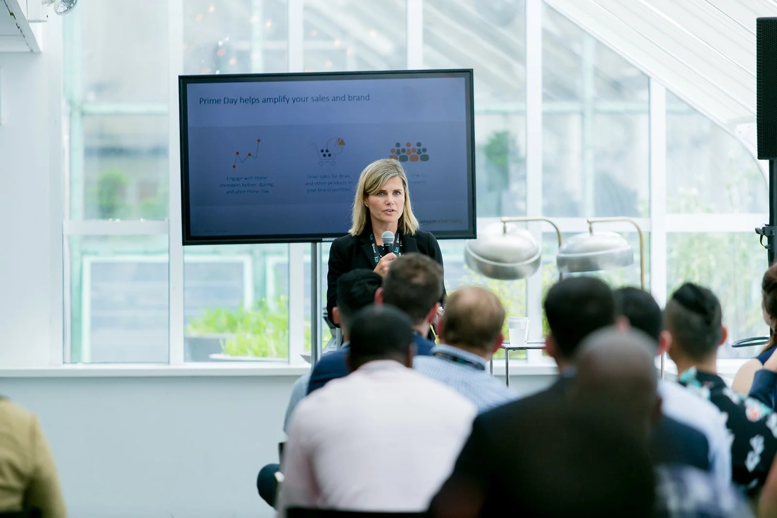 A woman in a black blazer giving a presentation to an audience in a bright, modern room with large windows. She is holding a microphone while standing in front of a large screen displaying a slide about Prime Day.