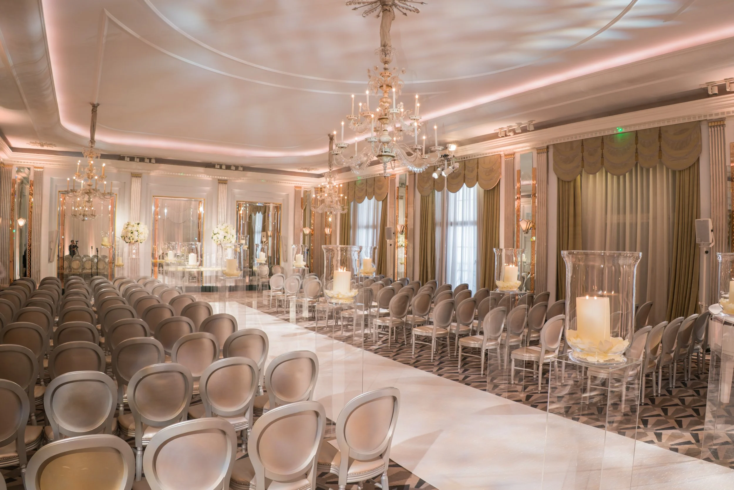 Elegant banquet hall decorated with chandeliers, candles, and gold curtains, set up for a formal event with rows of chairs facing each other.
