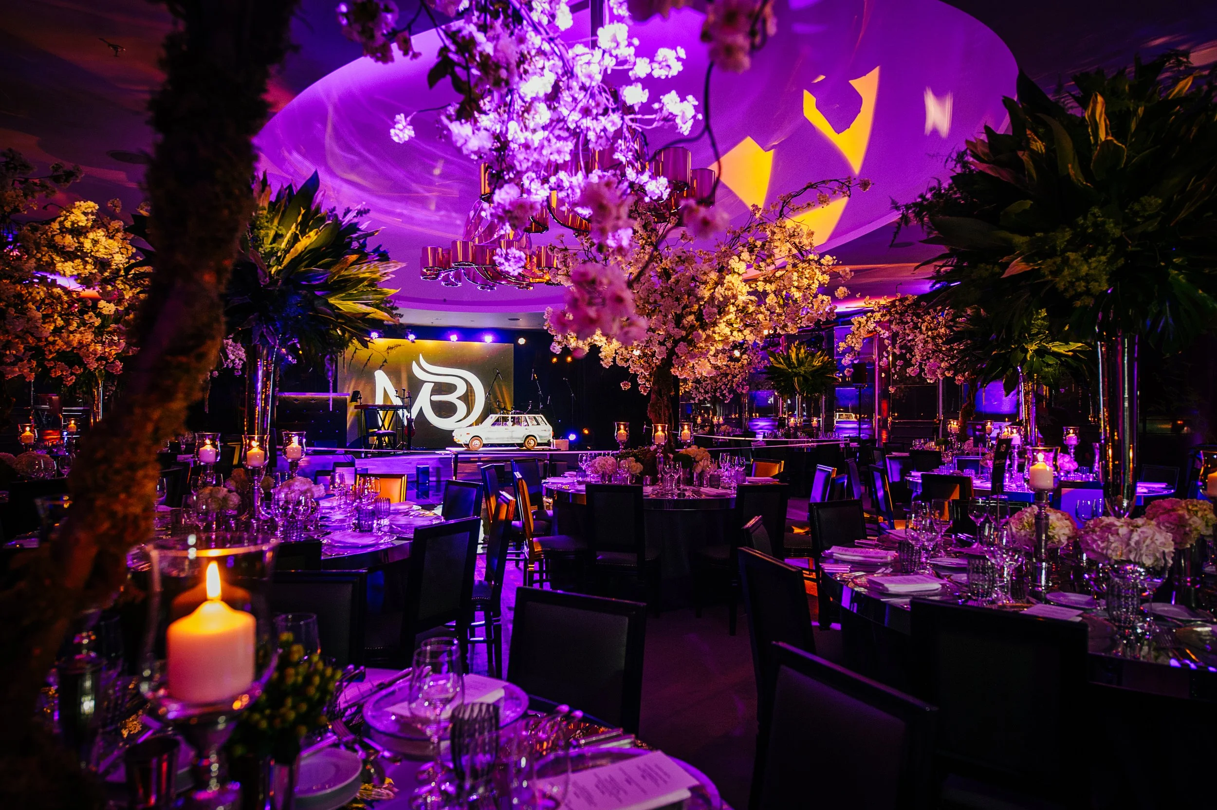 An event space decorated with pink and purple lighting, large floral arrangements, tables set with glassware and candles, and a small white vintage vehicle as a centerpiece. There are also banners or screens with a logo in the background.