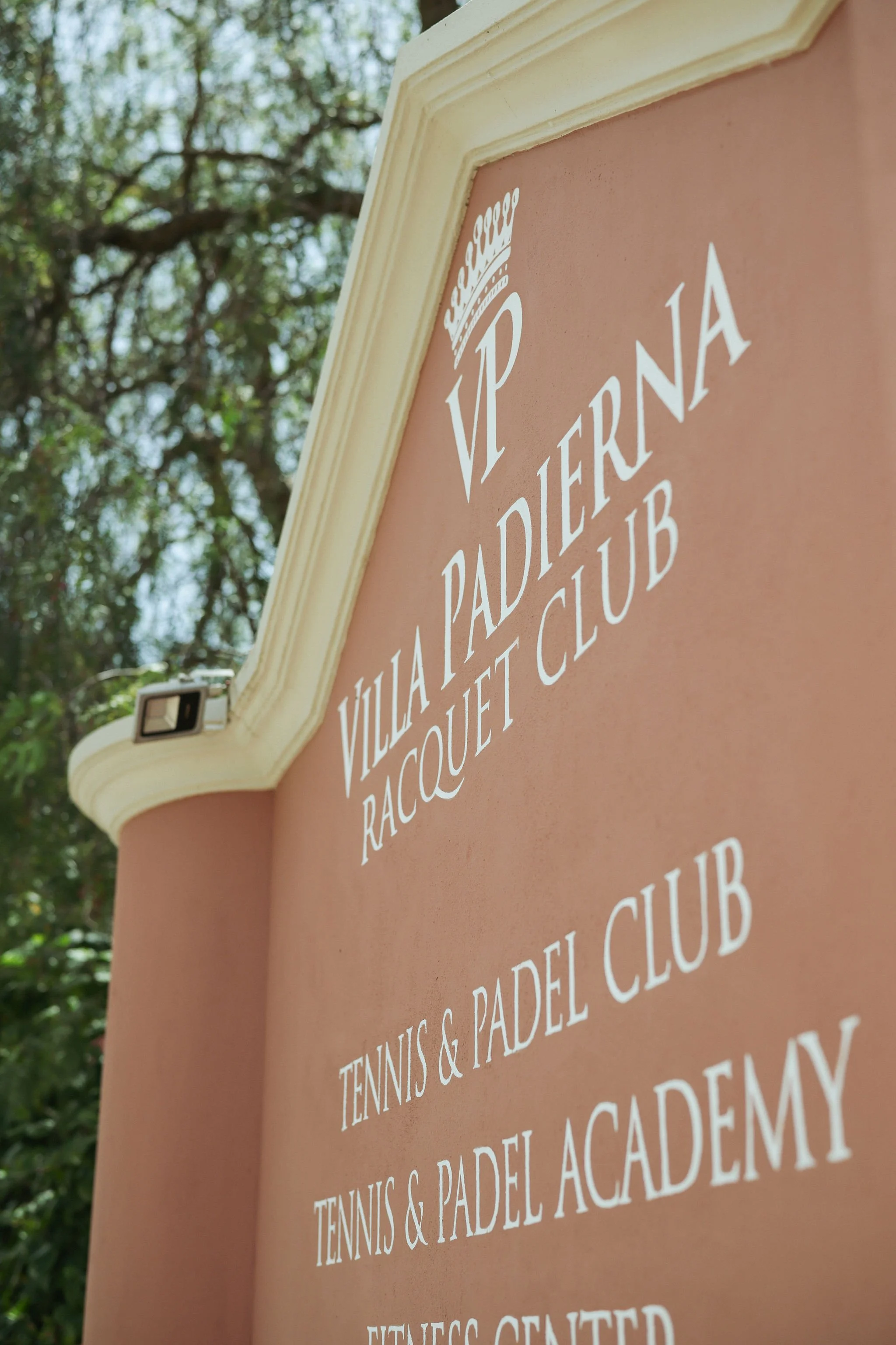 Close-up of a pink sign with white text reading "Vila Padella Tennis & Padel Club" on a beige border, outdoors with trees and sky in the background