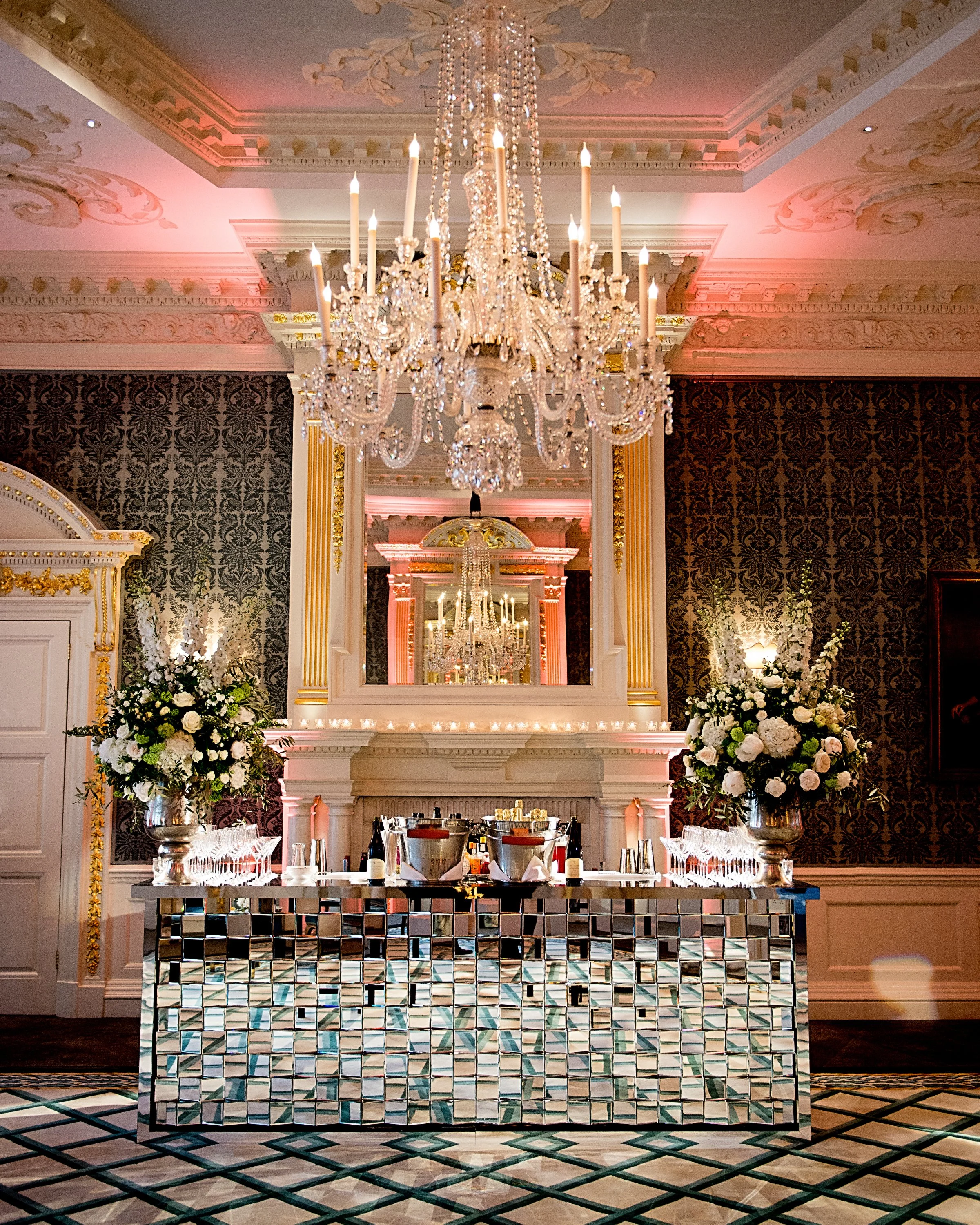 Elegant indoor setting with a large ornate chandelier hanging from a decorated ceiling, floral arrangements on either side of a mirrored bar with a geometric mirrored front, patterned wallpaper, and candle lights on a table.