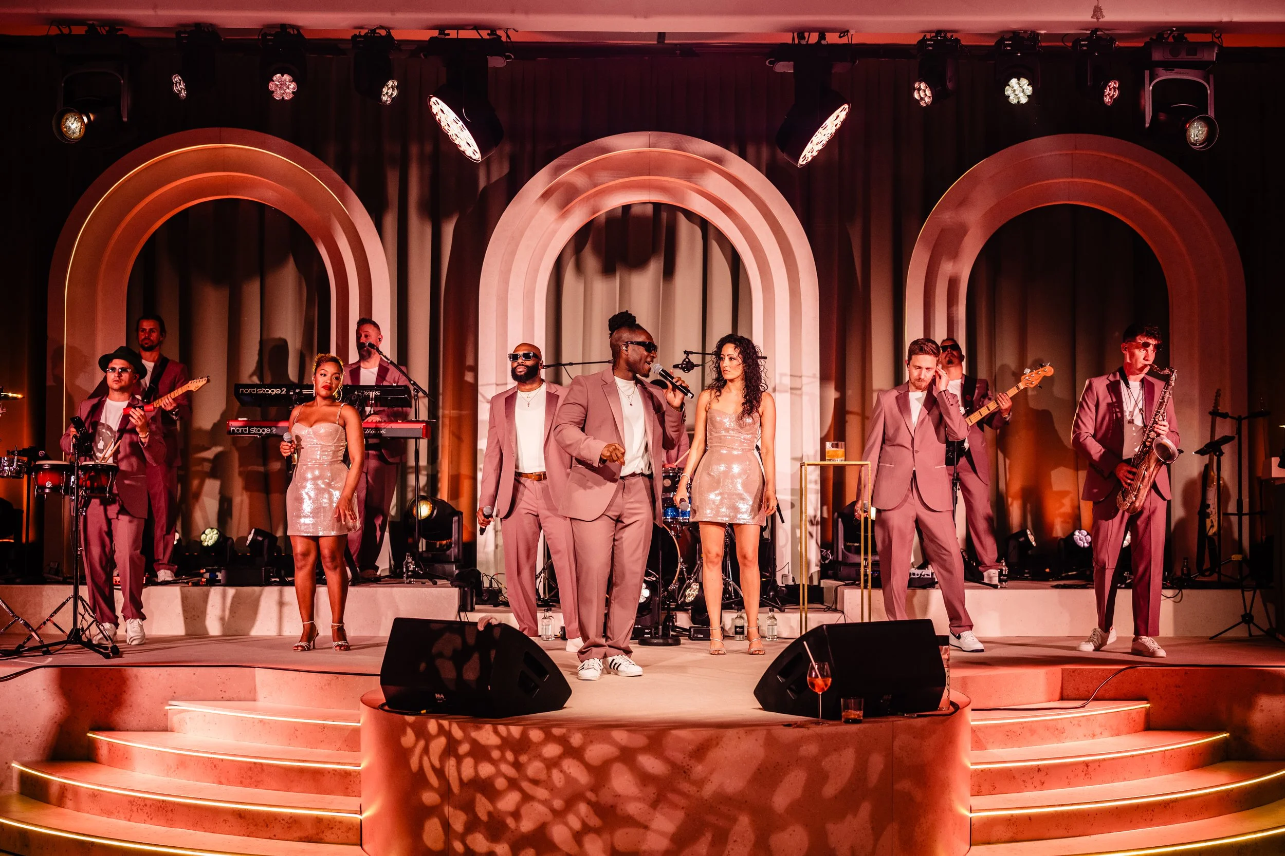 A band performing on stage with three women and six men, all dressed in matching pink suits, playing instruments and singing, under stage lighting with pink and orange hues at Fairmont Windsor Park'.