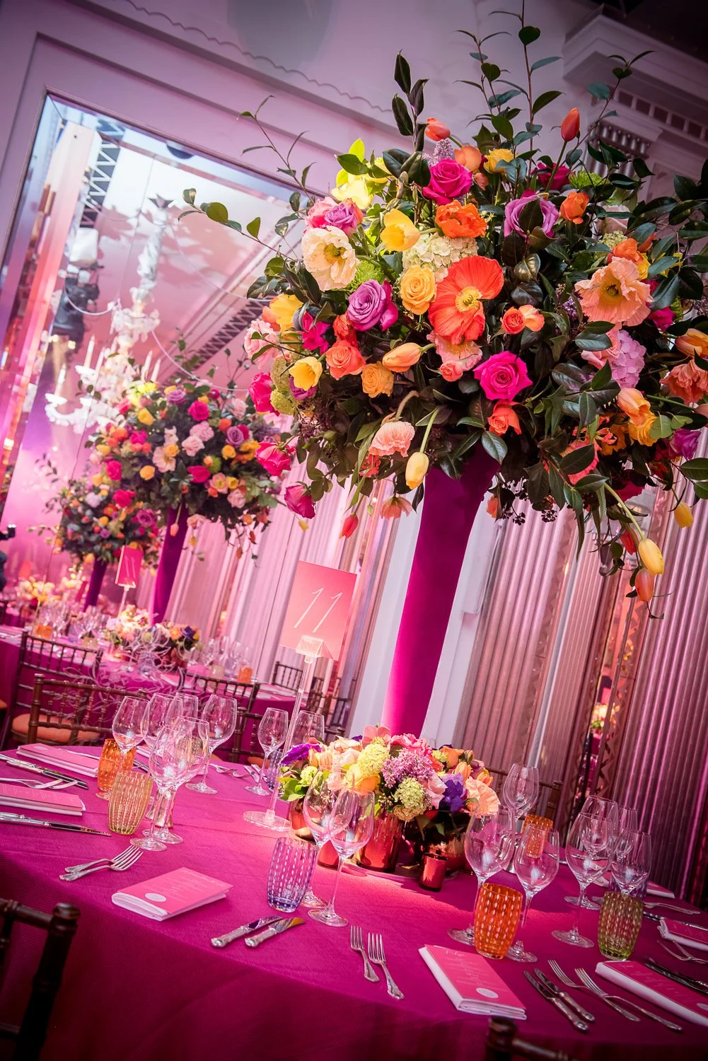 Elegant dining setup with pink tablecloths, tall floral centerpieces with pink, purple, yellow, and white flowers, and wine glasses in a decorated venue with pink lighting.