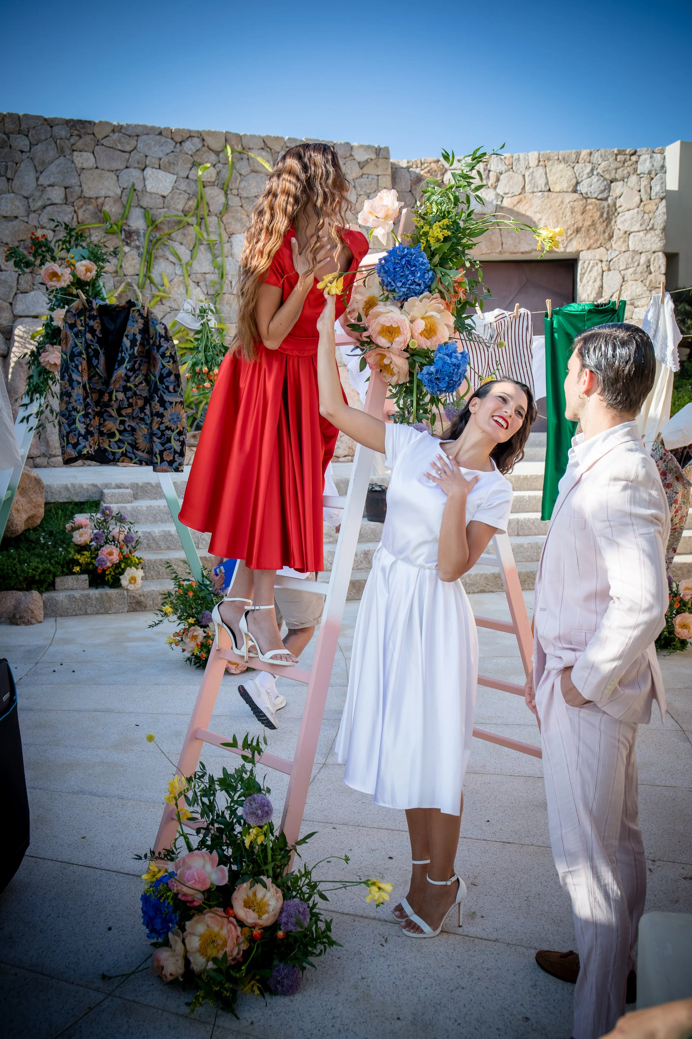 A woman in a red dress standing on a ladder with another woman in a white dress, while a man kneels with his hand on her shoulder, all surrounded by floral decorations and clothes hanging on a line, outdoors near a stone wall.