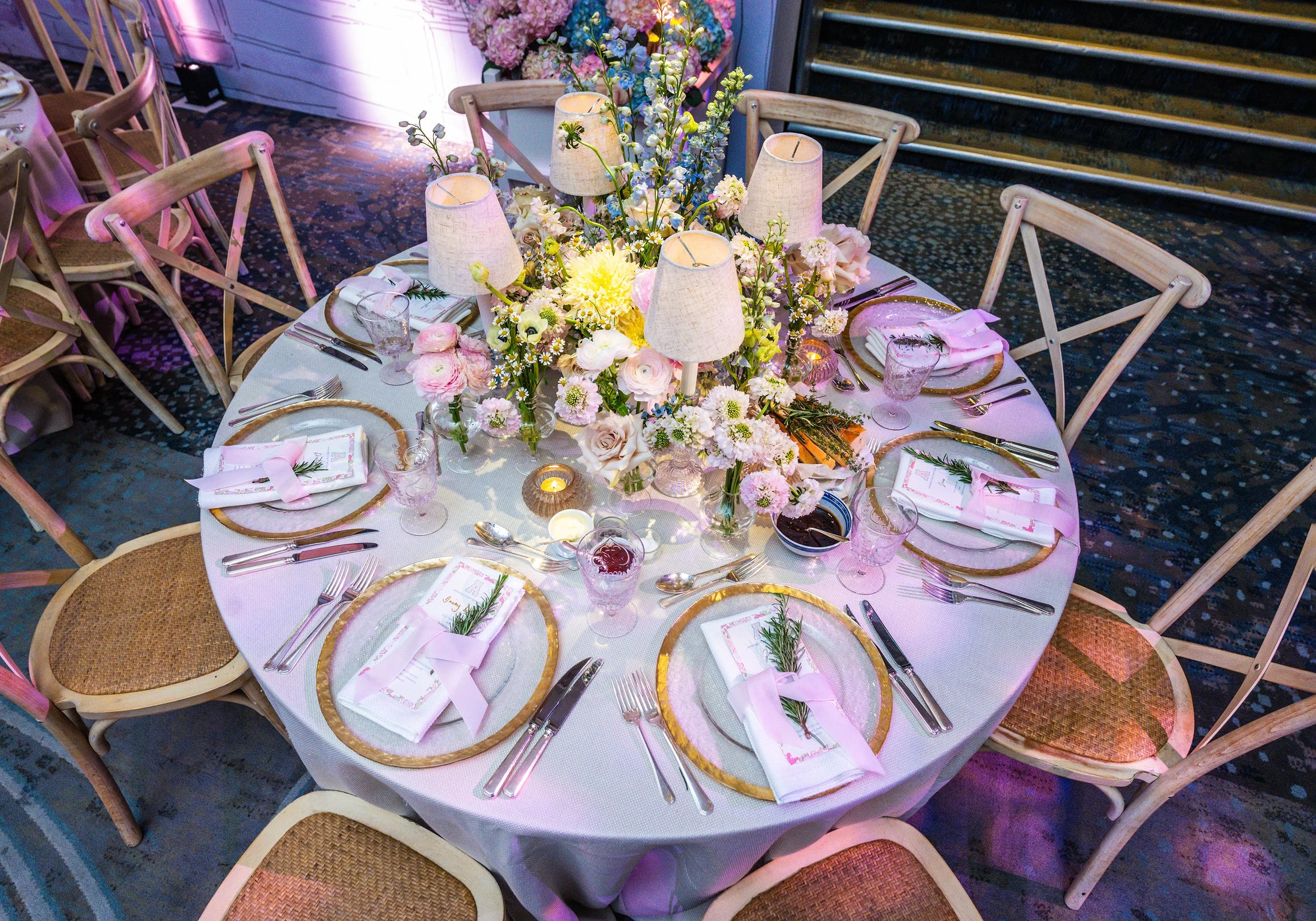 A round banquet table decorated with pink and white flowers, small lamps, and gold-rimmed plates, set for a Bat Mitzvah at Grosvenor House on Park Lane 