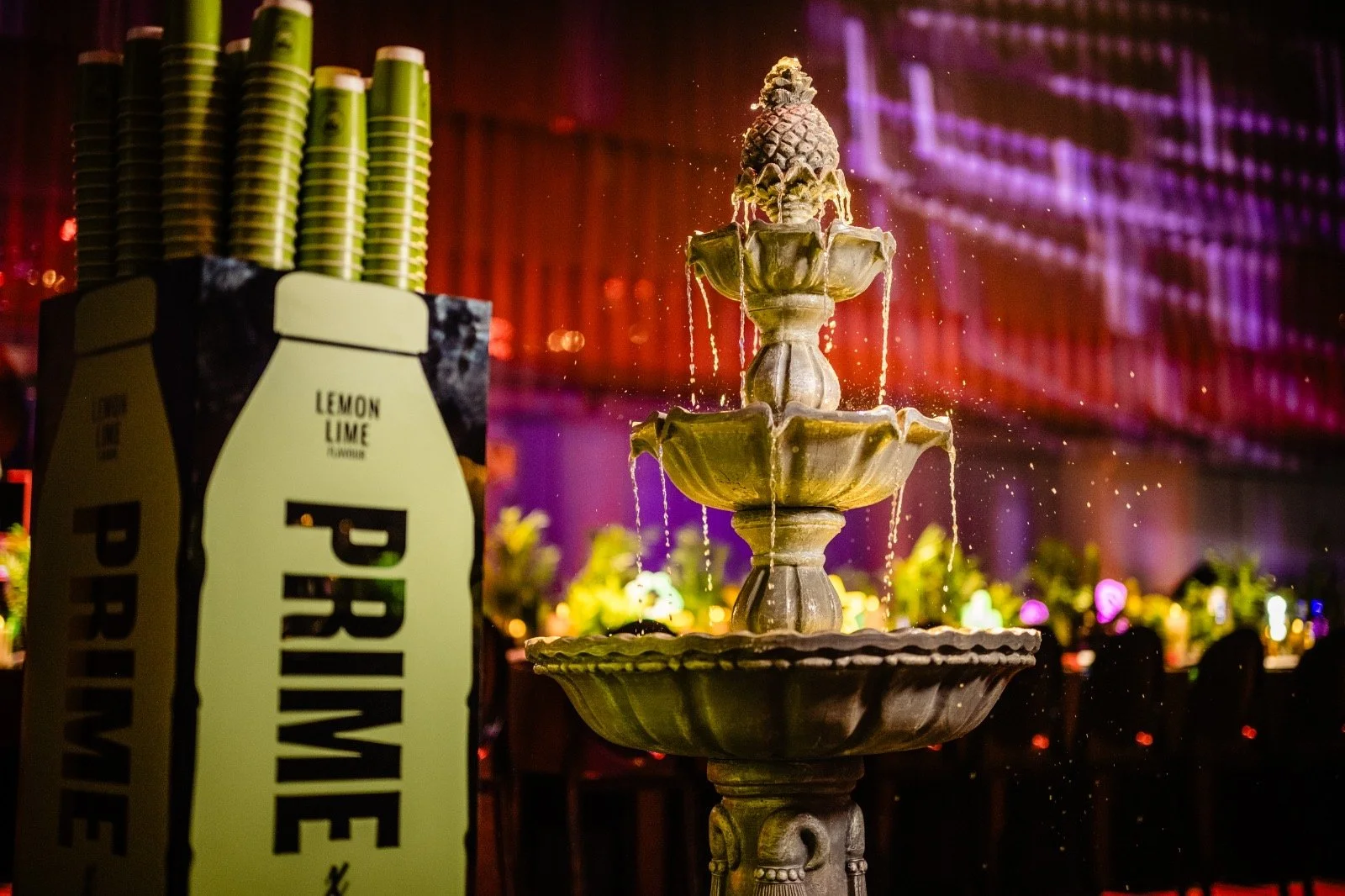 A decorative fountain with a pineapple-shaped top in a dimly lit setting. To the left, a box labeled "PRIME" with yellow cups on top. The background features colorful lighting and tables with flowers, suggesting a party or banquet environment 