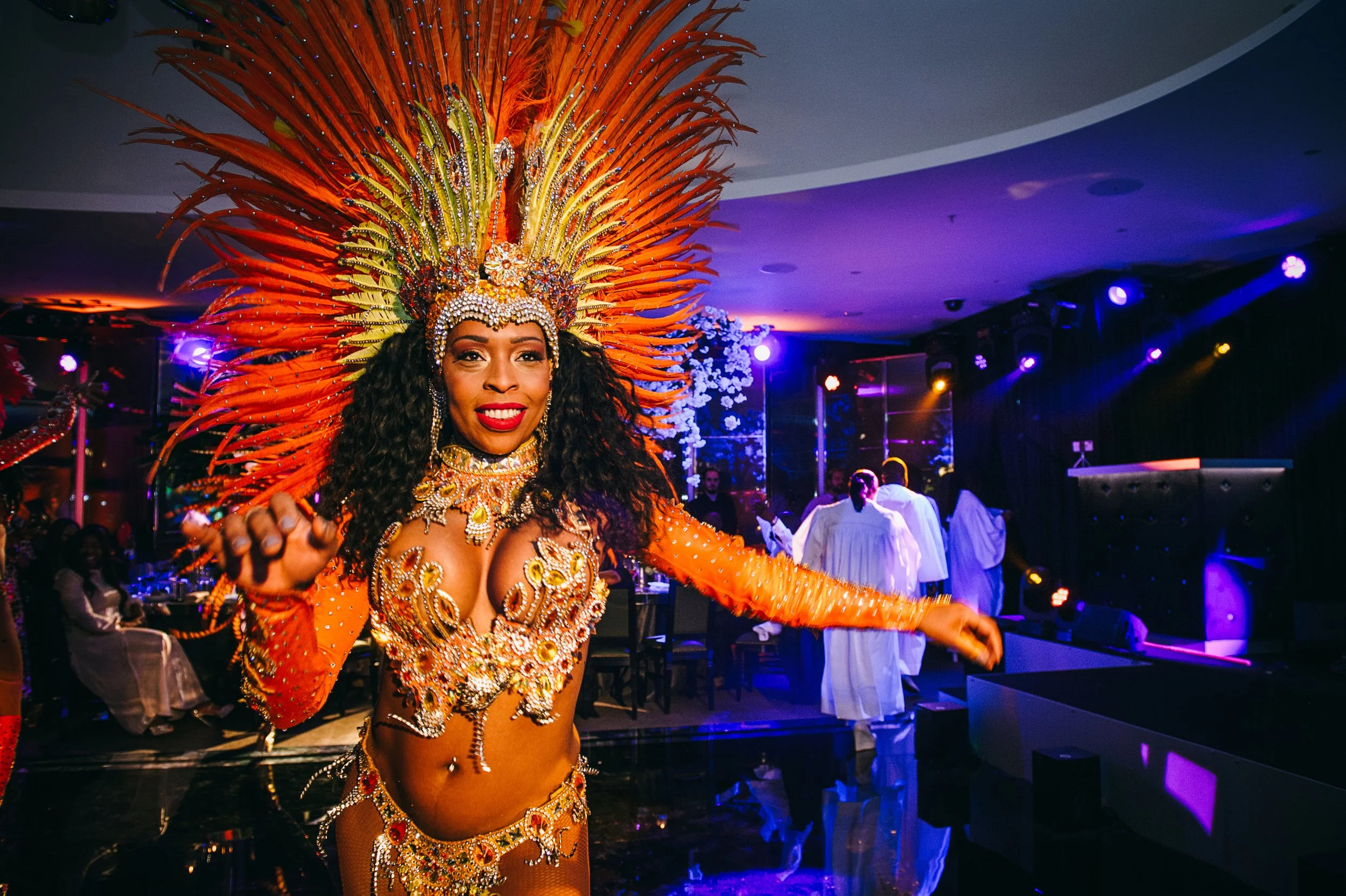 A woman dressed in a colorful, elaborate carnival costume with a large feathered headdress, standing and smiling at a lively indoor event with purple and blue lighting.