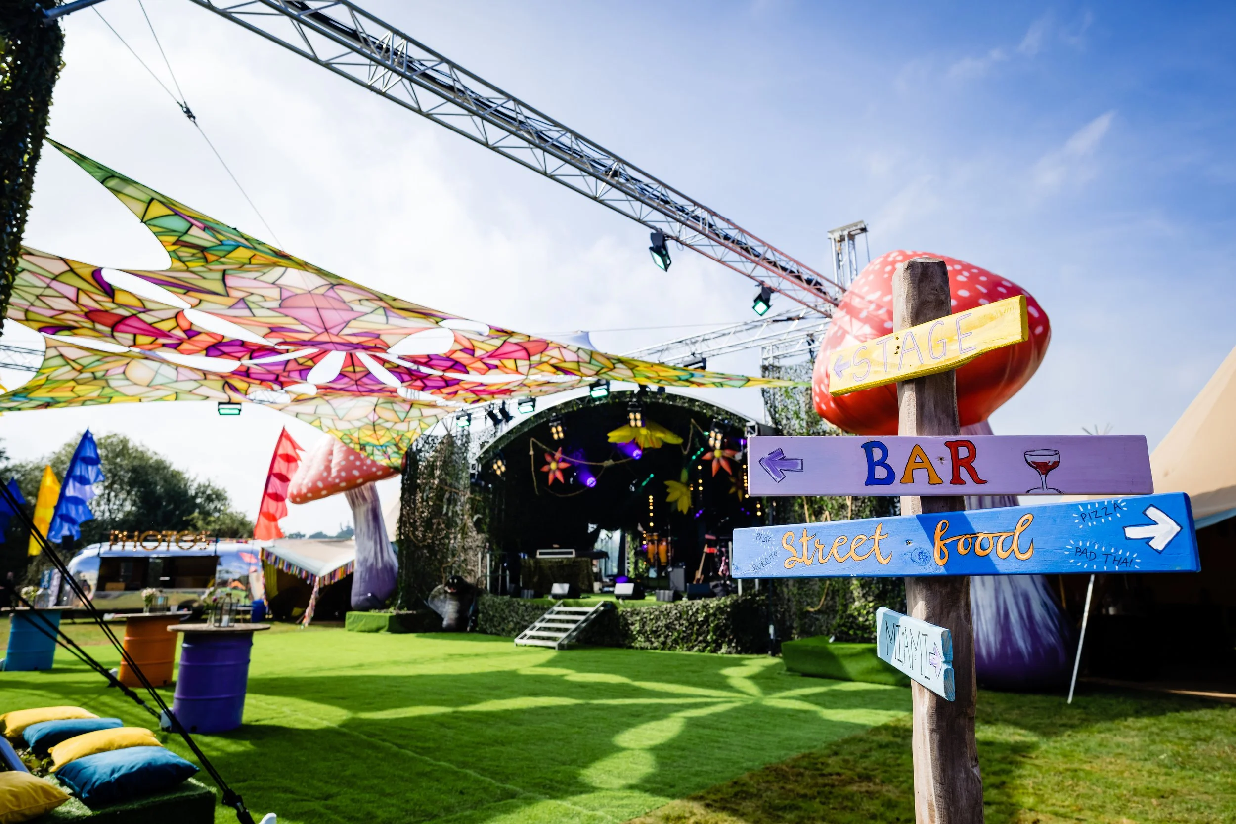 Colorful street festival with decorative canopy, food and drink signs, stage, and large mushroom-shaped sculpture under a partly cloudy sky at Bar Mitzvah at  Wrotham Park