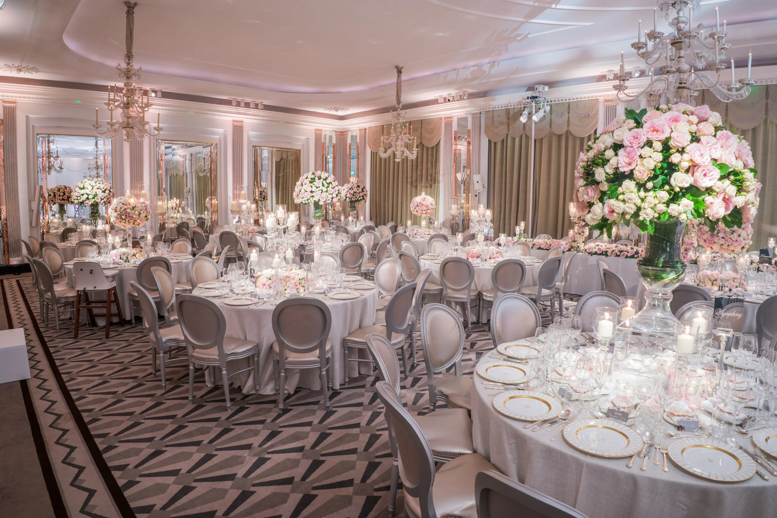 Elegant banquet hall decorated for a wedding reception with round tables covered in white tablecloths, floral centerpieces, china, glassware, and candles, surrounded by cream-colored chairs, ornate chandeliers, tall curtains, and mirrors on the walls
