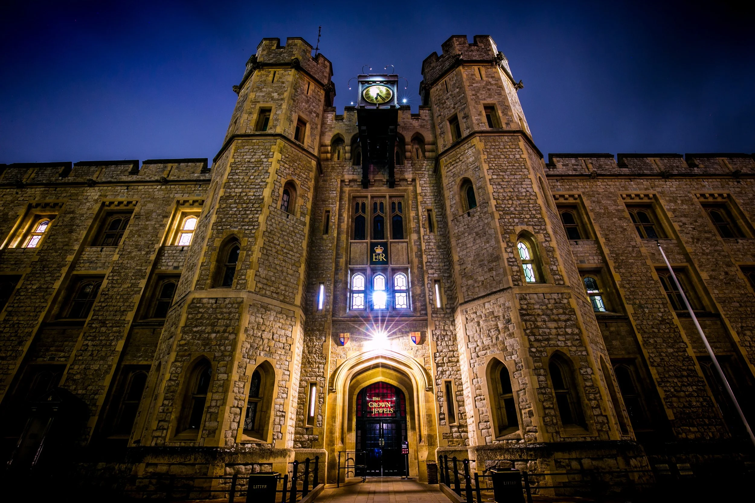 Night view of a historic stone castle-like building with a clock tower, illuminated windows, and a glowing sign that reads 'The Crown Jewels' at the entrance.