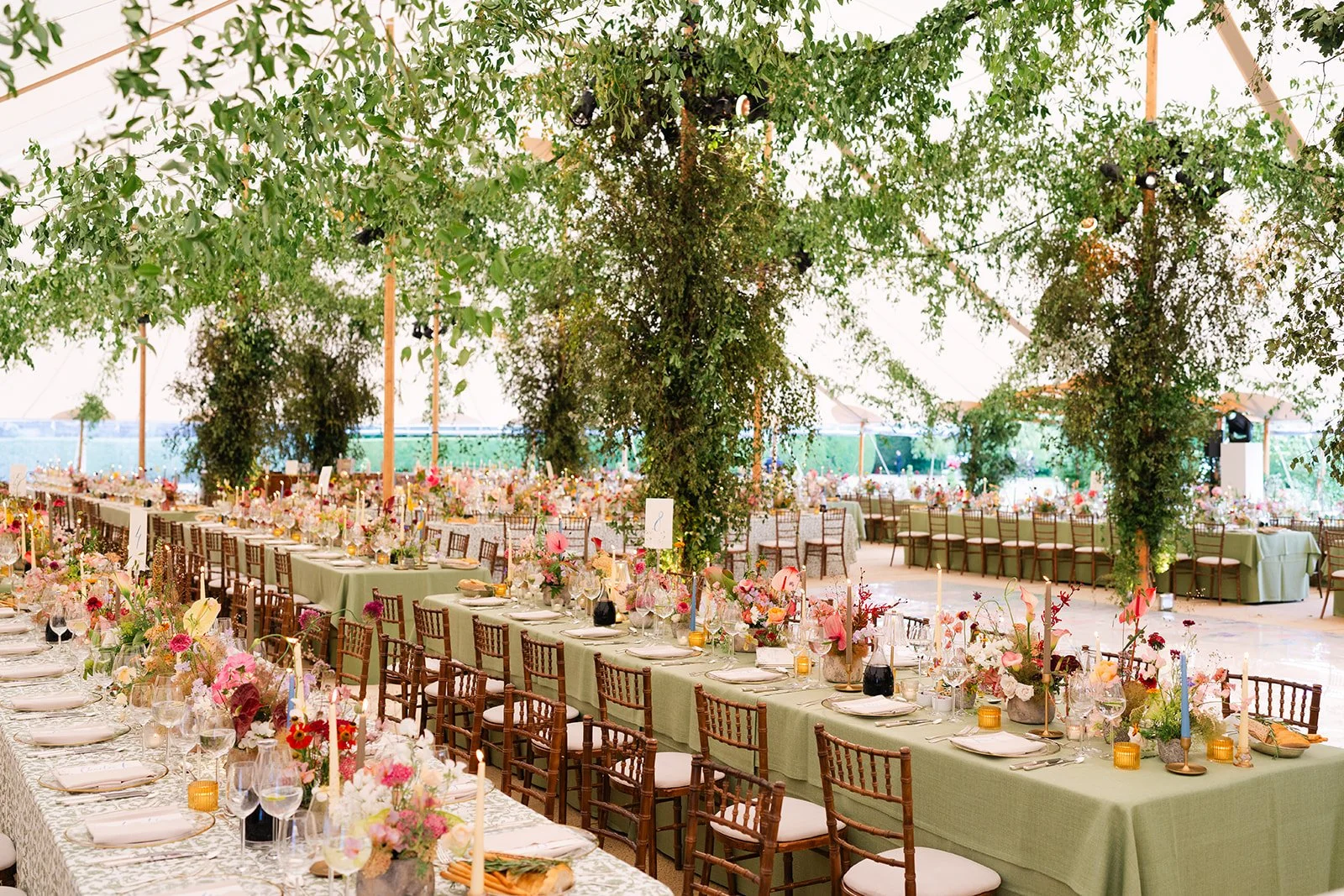 An elegant weddding decorated with long tables covered in green tablecloths, set with plates, glasses, and floral centerpieces, surrounded by chairs, with a ceiling of green foliage and hanging lights.