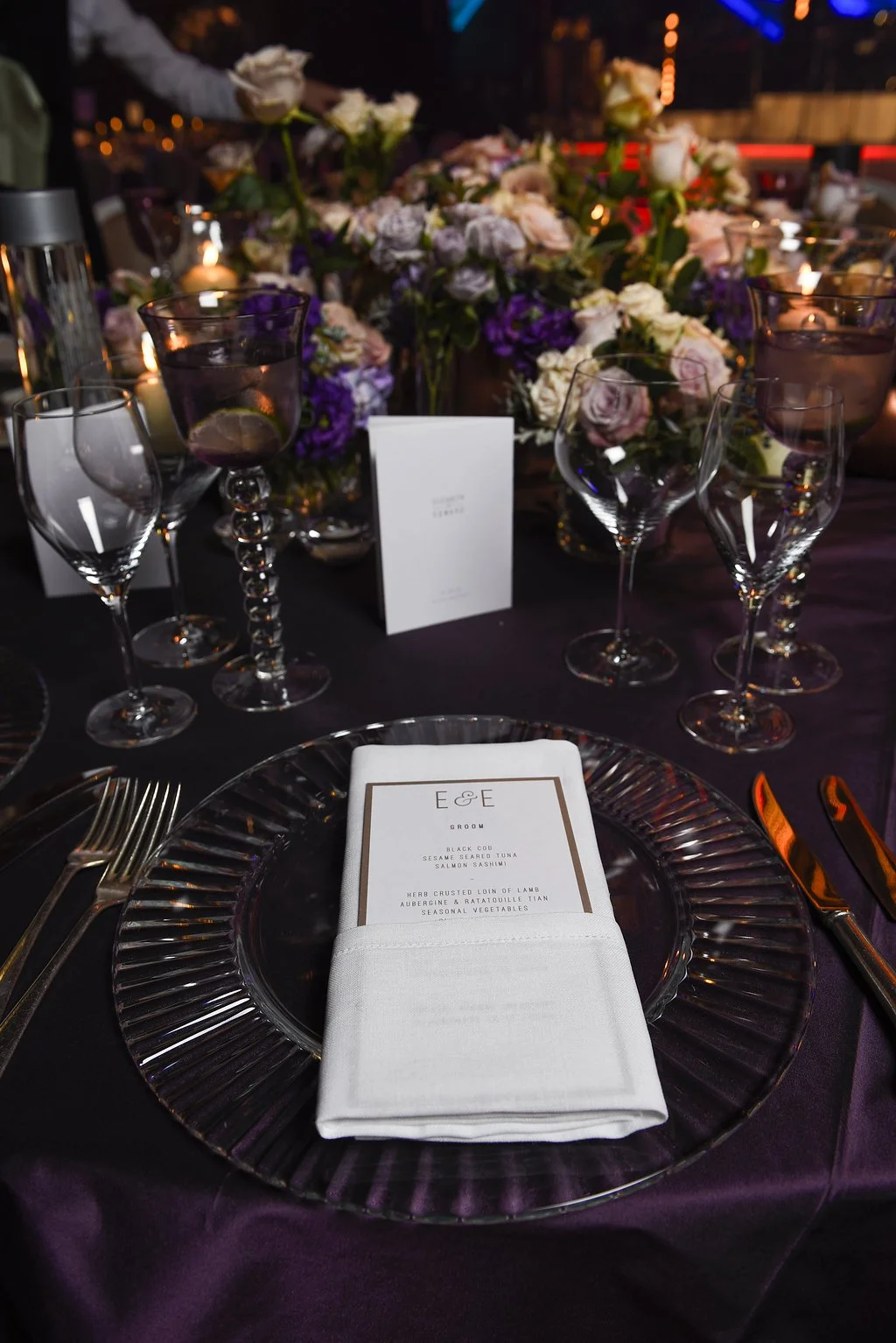 Elegant table setting with a glass charger plate, gold flatware, a folded white napkin with a printed menu, multiple wine glasses, and a centerpiece of pink and purple flowers and candles in a dimly lit setting.