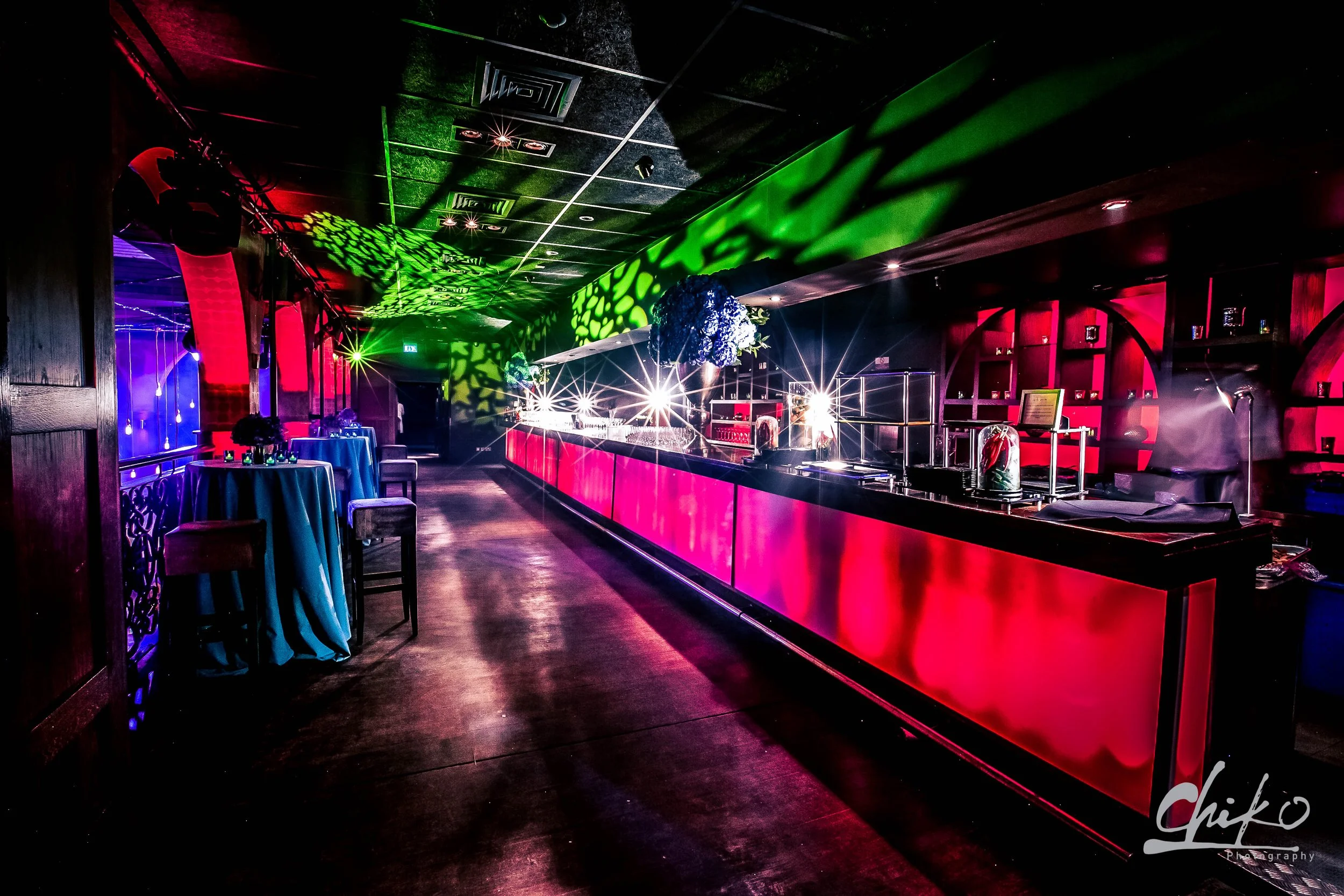 A dimly lit nightclub interior with a red bar counter, purple and green lighting, tables with blue tablecloths, and decorative floral arrangements hanging above. Bright lasers and spotlights create colorful patterns on the ceiling.