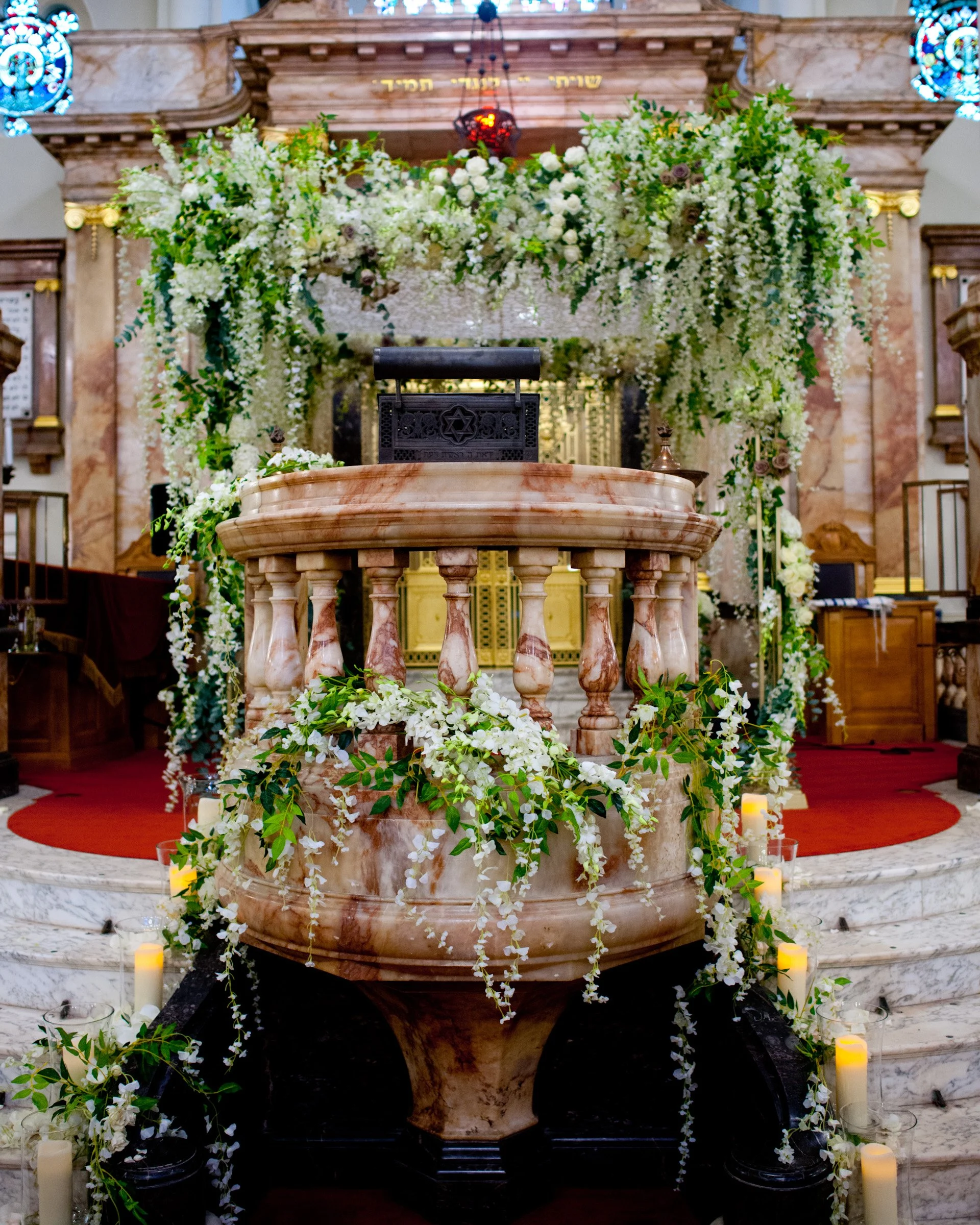 A floral arrangement surrounds a marble baptismal font in a church, with candles and greenery on the floor. The altar behind is decorated with more flowers and woodwork.