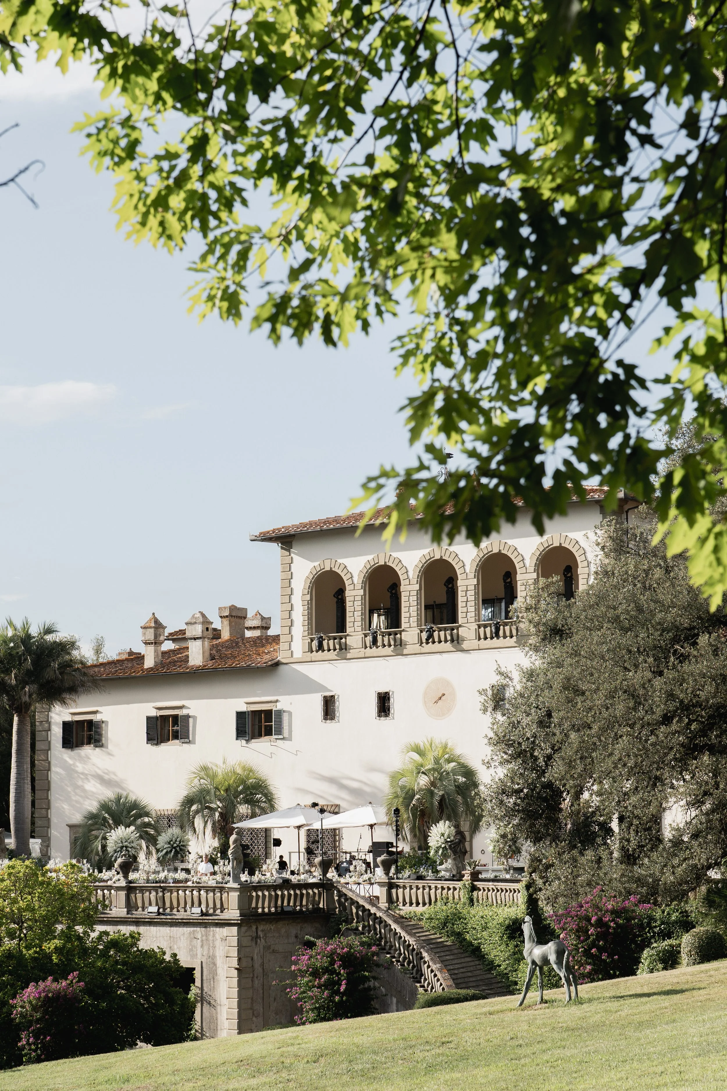 A wdding location in Florence called Villa Palmieri which is a grand white building with arches, surrounded by lush greenery, trees, and flowers, with a sculpture of a horse in the foreground.