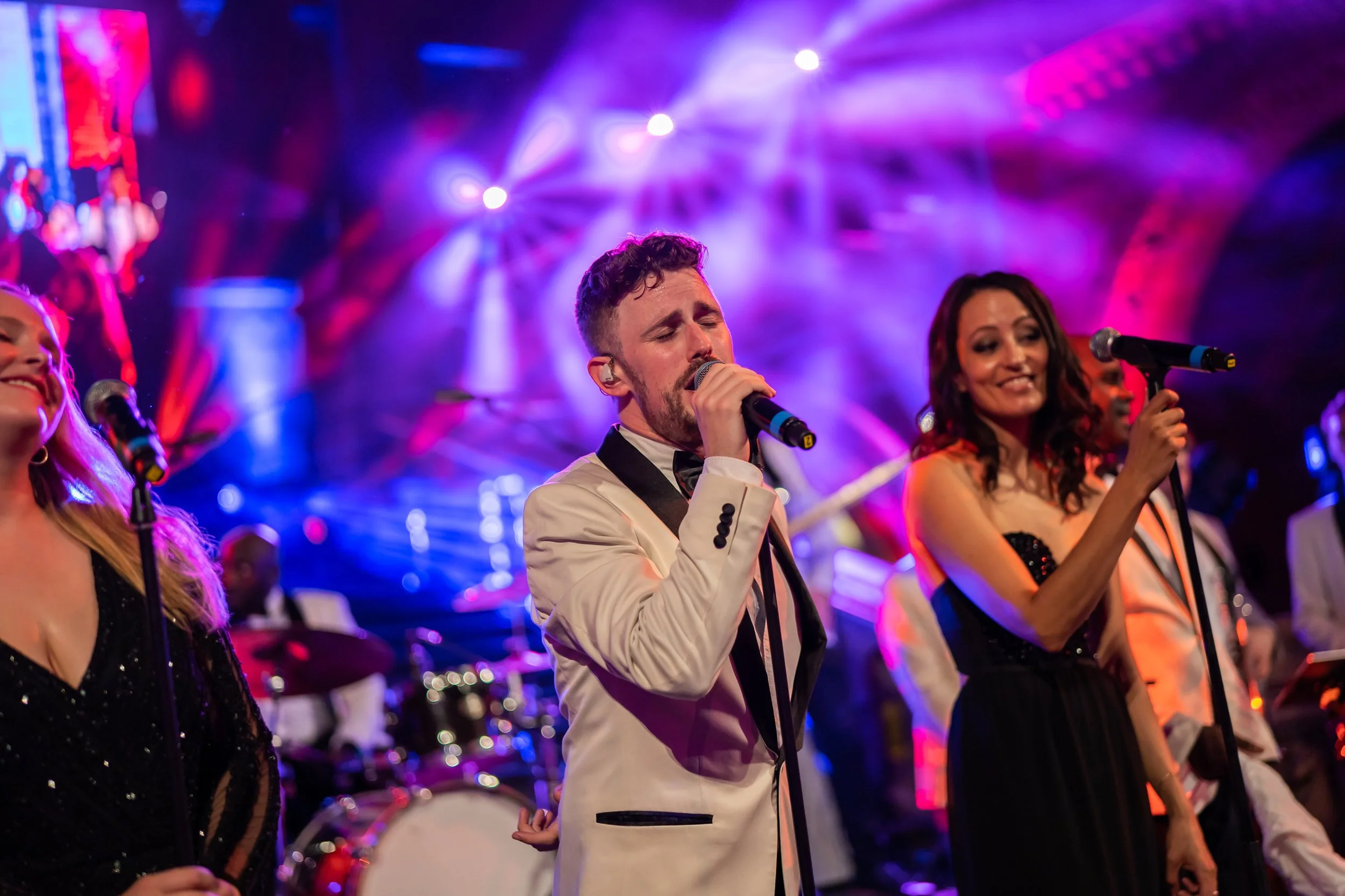 A male singer with curly hair and a beard in a white suit and black bow tie singing passionately on stage with colorful purple and pink lights behind him at The Natural History Museum