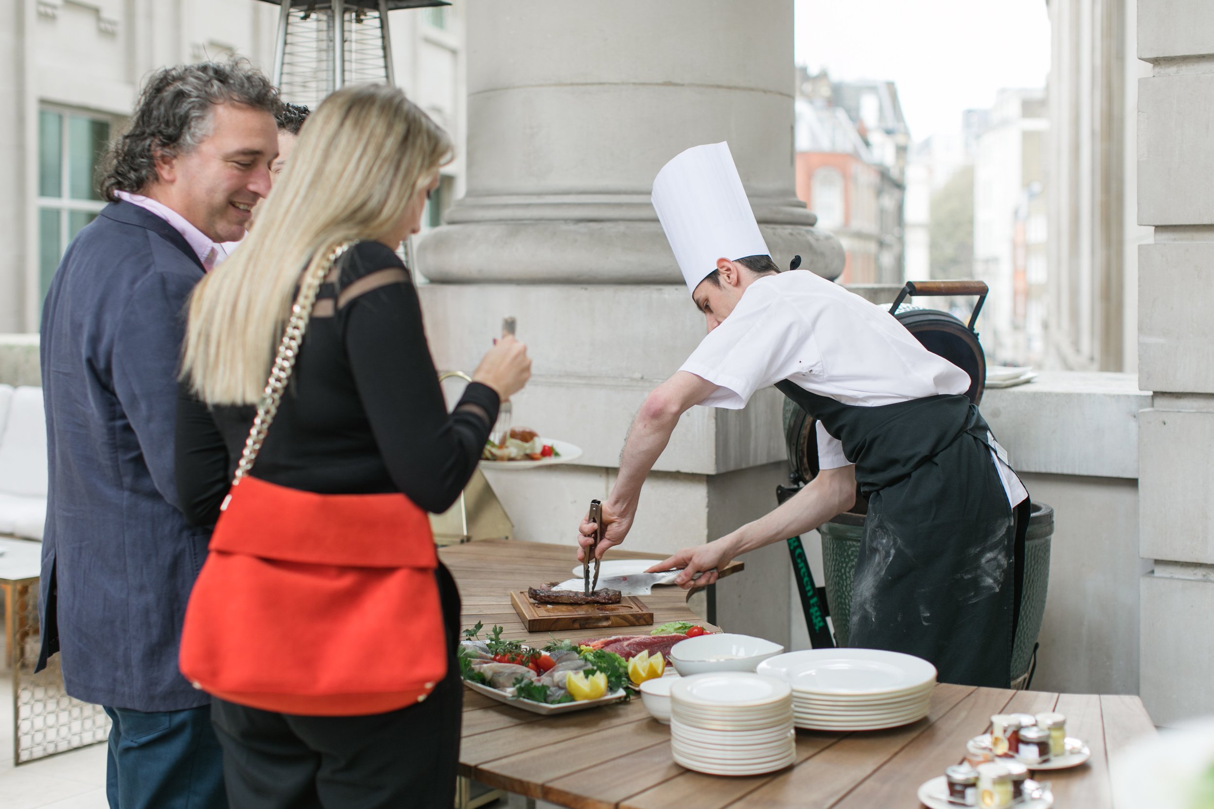 A chef slicing grilled meat at an outdoor buffet with a woman and a man serving themselves food.
