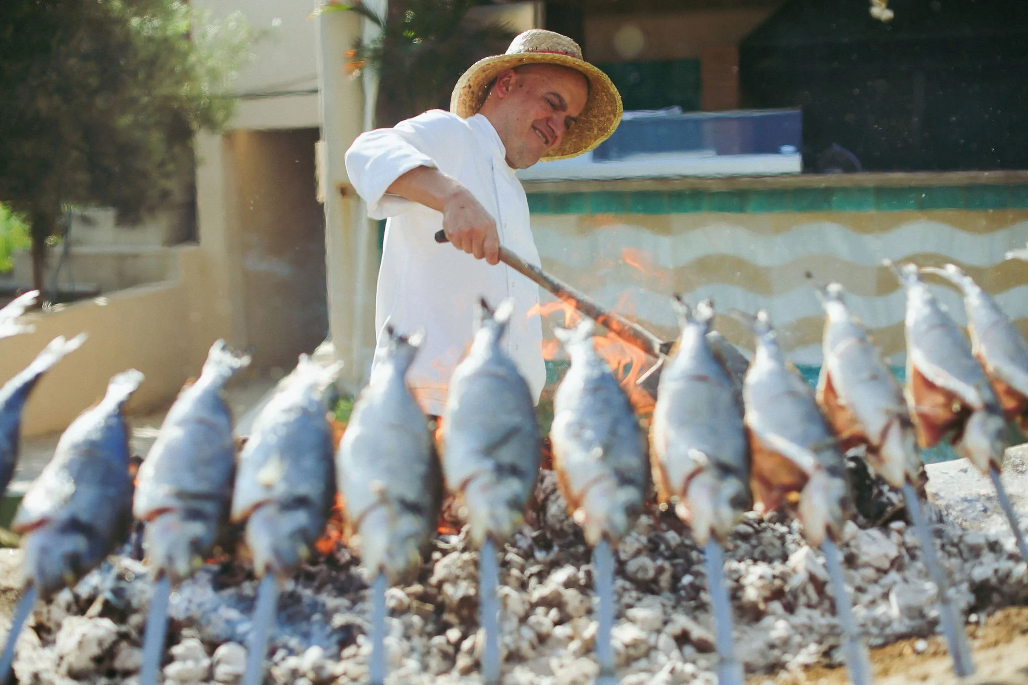 A man wearing a straw hat and white shirt turns while grilling fish over an open flame outside at Anantara Villa Padierna Resort in Marbella