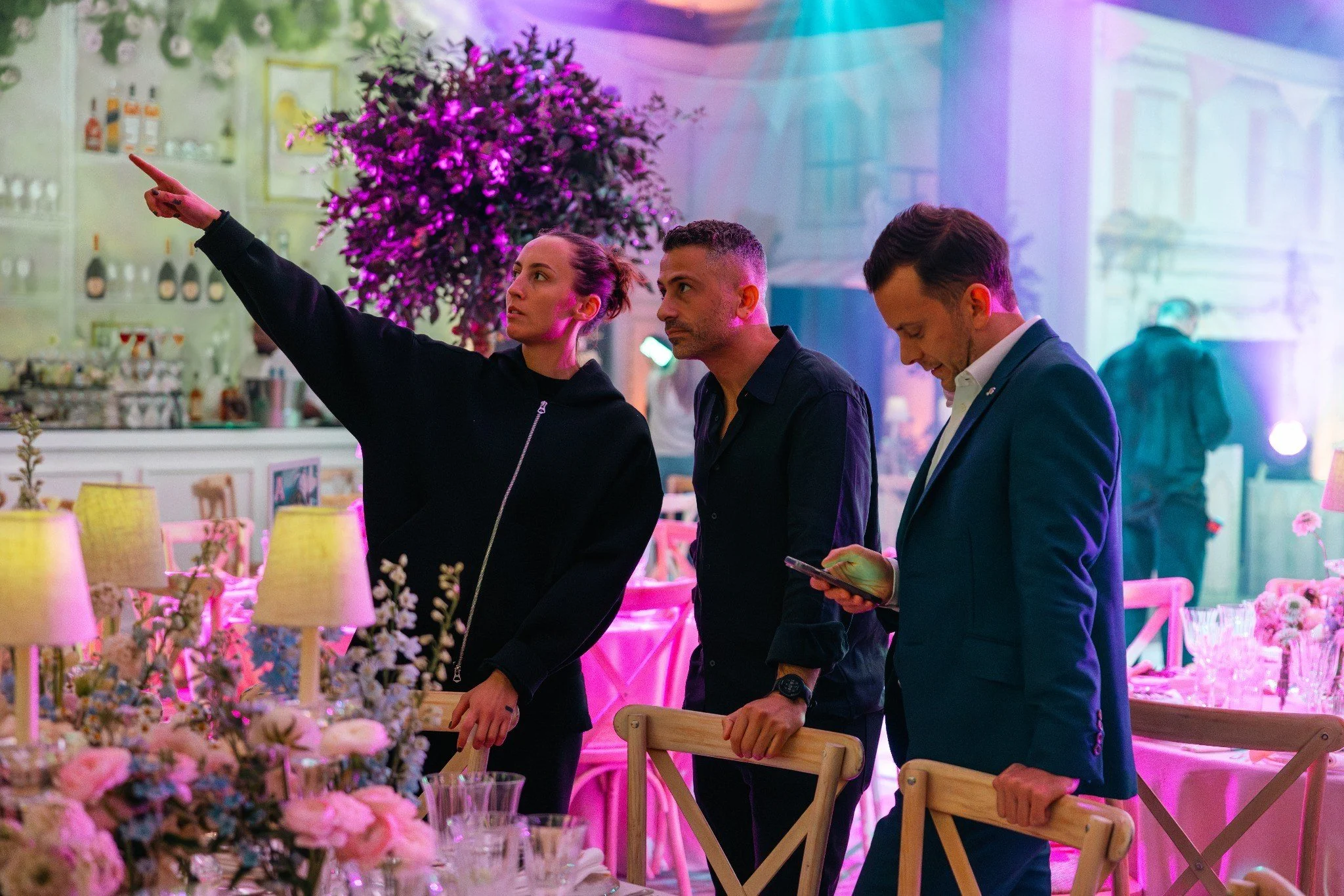 Setting up Hamptons themed Bat Mitzvah A woman with her hair in a bun pointing with her left hand while talking to two men at a colorful event with purple and blue lighting and decorated with flowers.