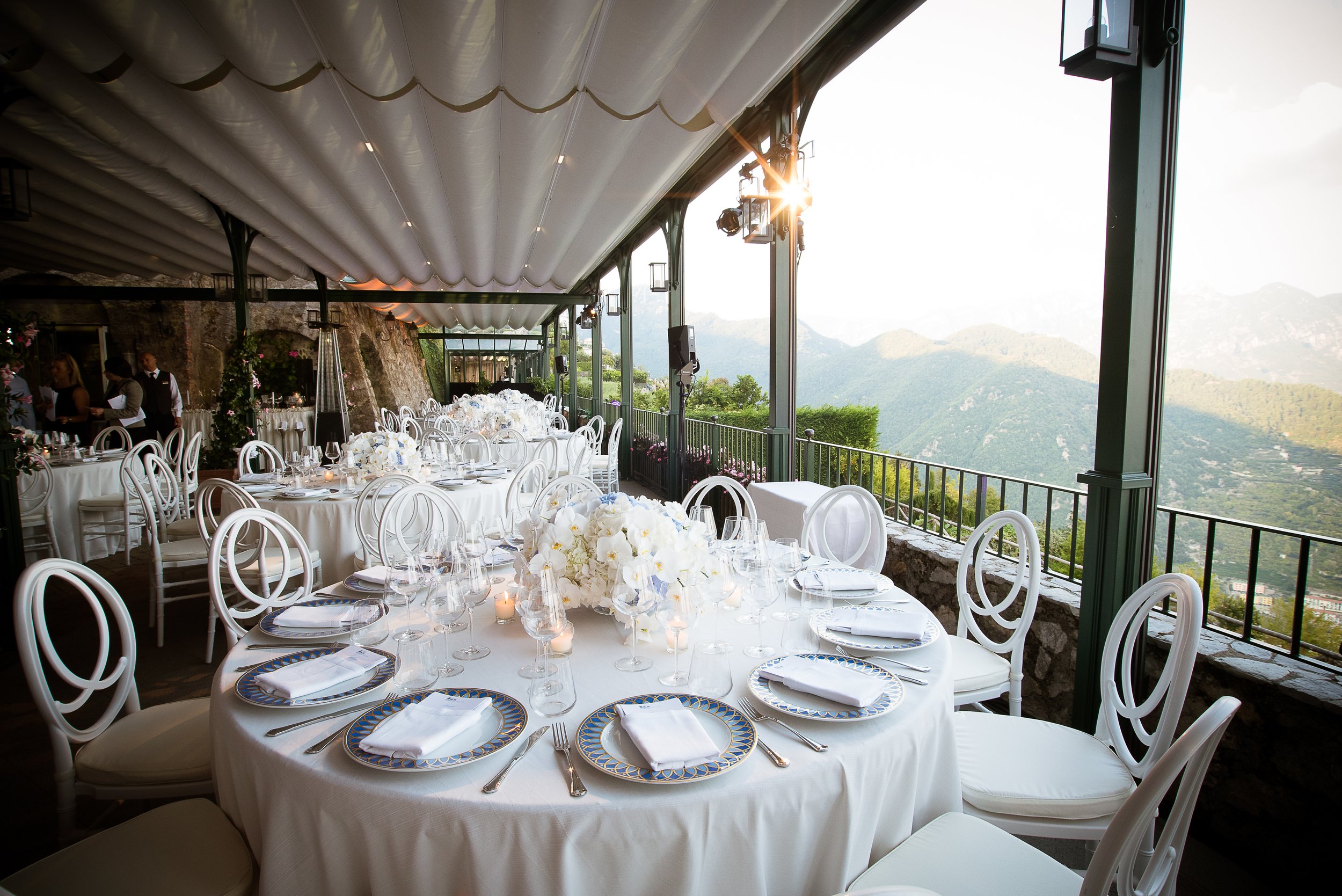 A beautifully decorated outdoor wedding reception area with white round tables, floral centerpieces, and elegant chairs, situated on a terrace with a scenic mountain view and a setting sun.